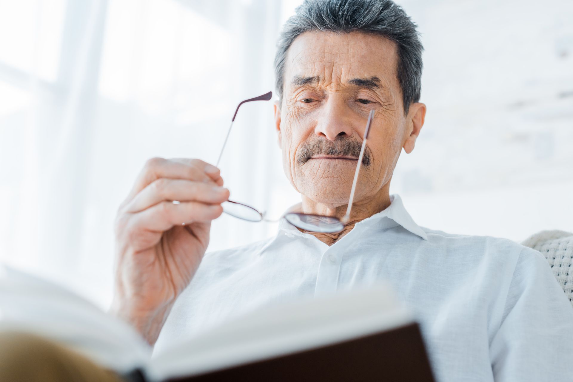Elderly man reading a book, holding glasses. He is wearing a white shirt and has a mustache, in a bright room.