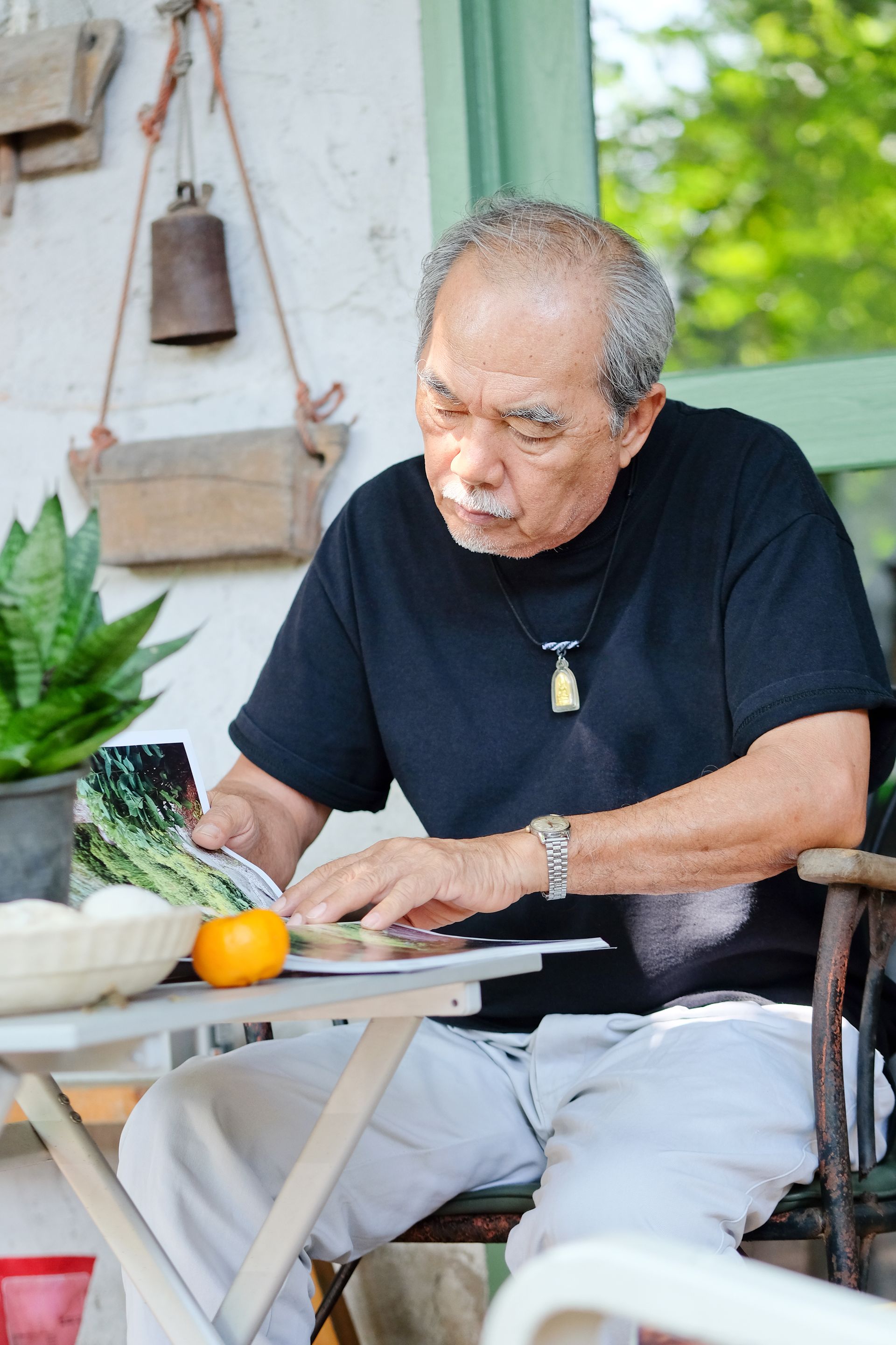 Elderly man with gray hair, in black shirt, examining object at table outdoors.