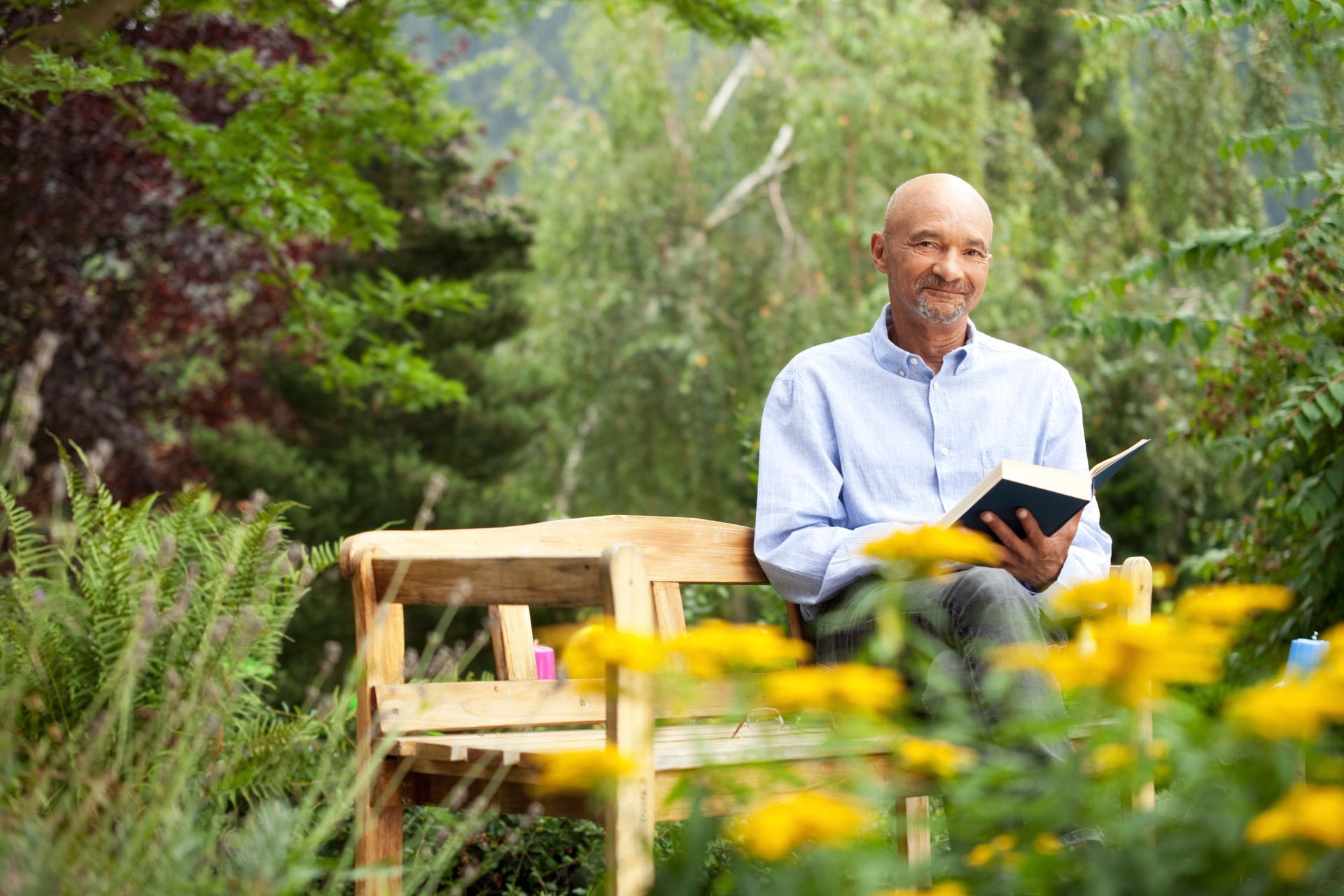 Bald man reading a book on a wooden bench in a garden with flowers and trees, smiling at the camera.