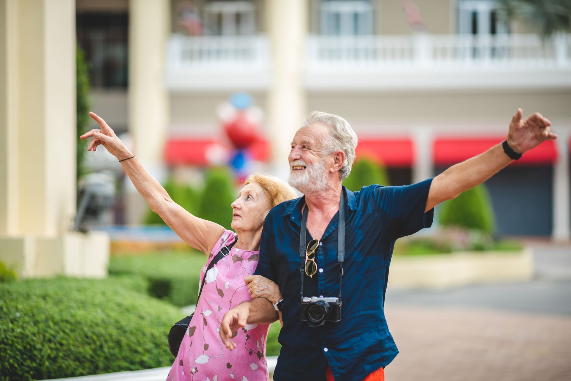 Elderly couple walking outdoors, arms up, looking and smiling. Man with camera around neck.