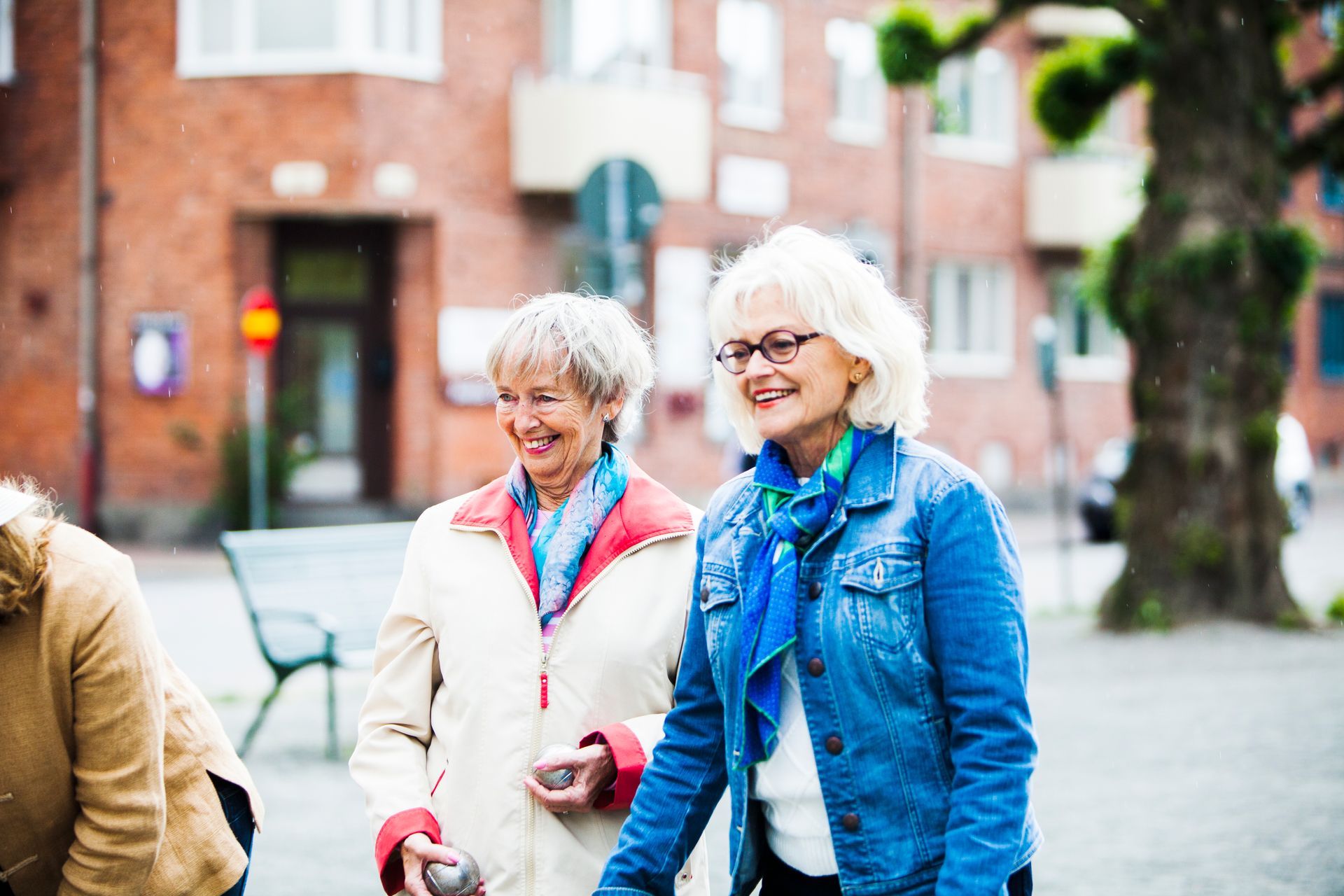 Two smiling elderly women playing boules outdoors near a brick building.