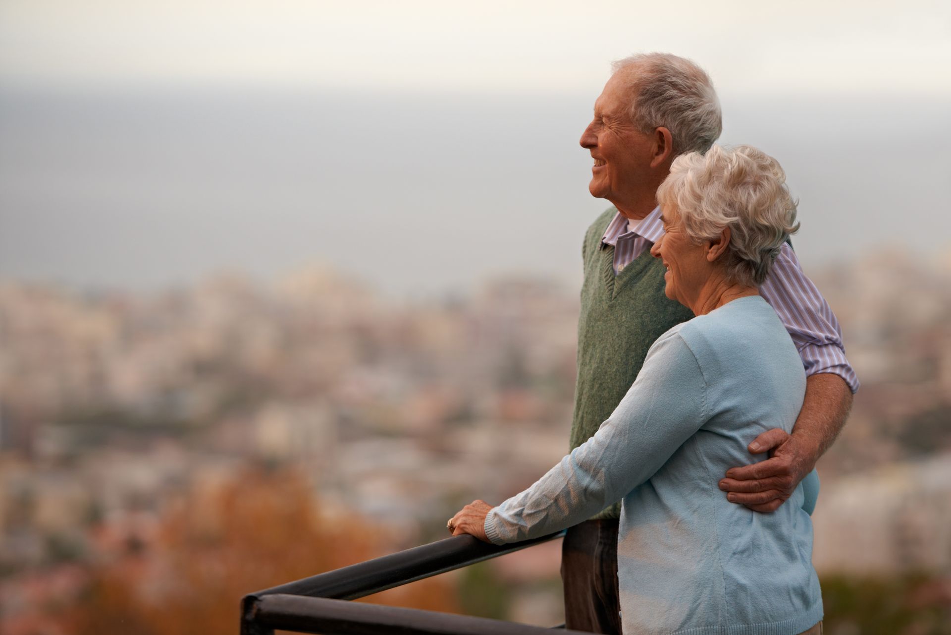 Elderly couple with gray hair smiles, embracing on a balcony overlooking a city.