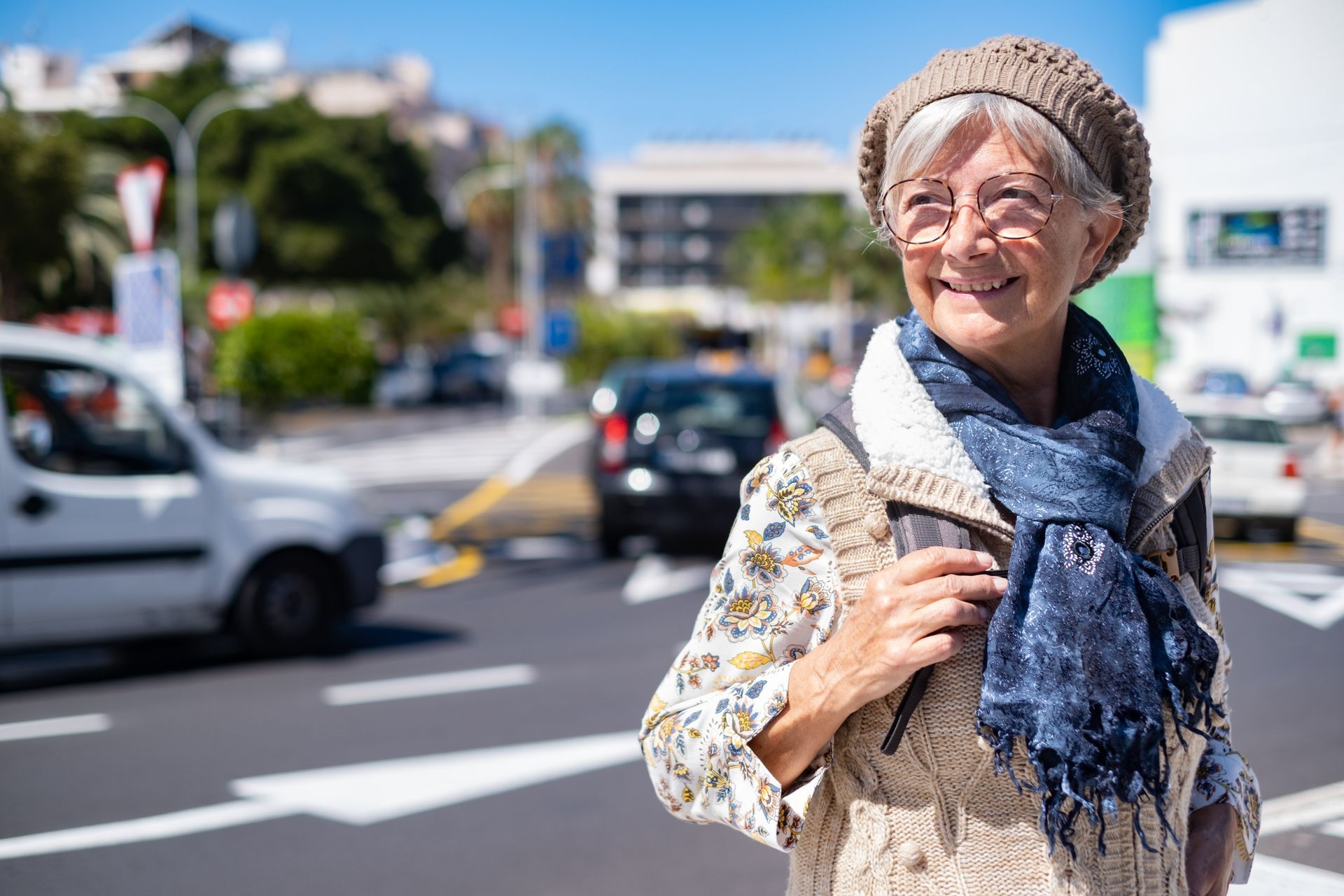 Elderly woman in a knitted hat smiles on a city street, holding a phone and looking to the side.