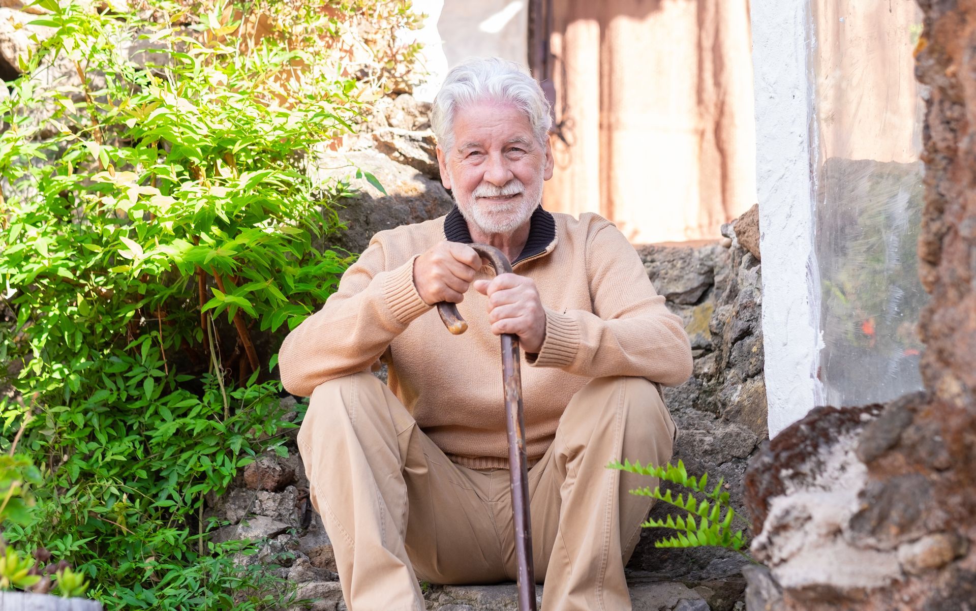Elderly man with white beard and cane sits on stone steps, smiling.