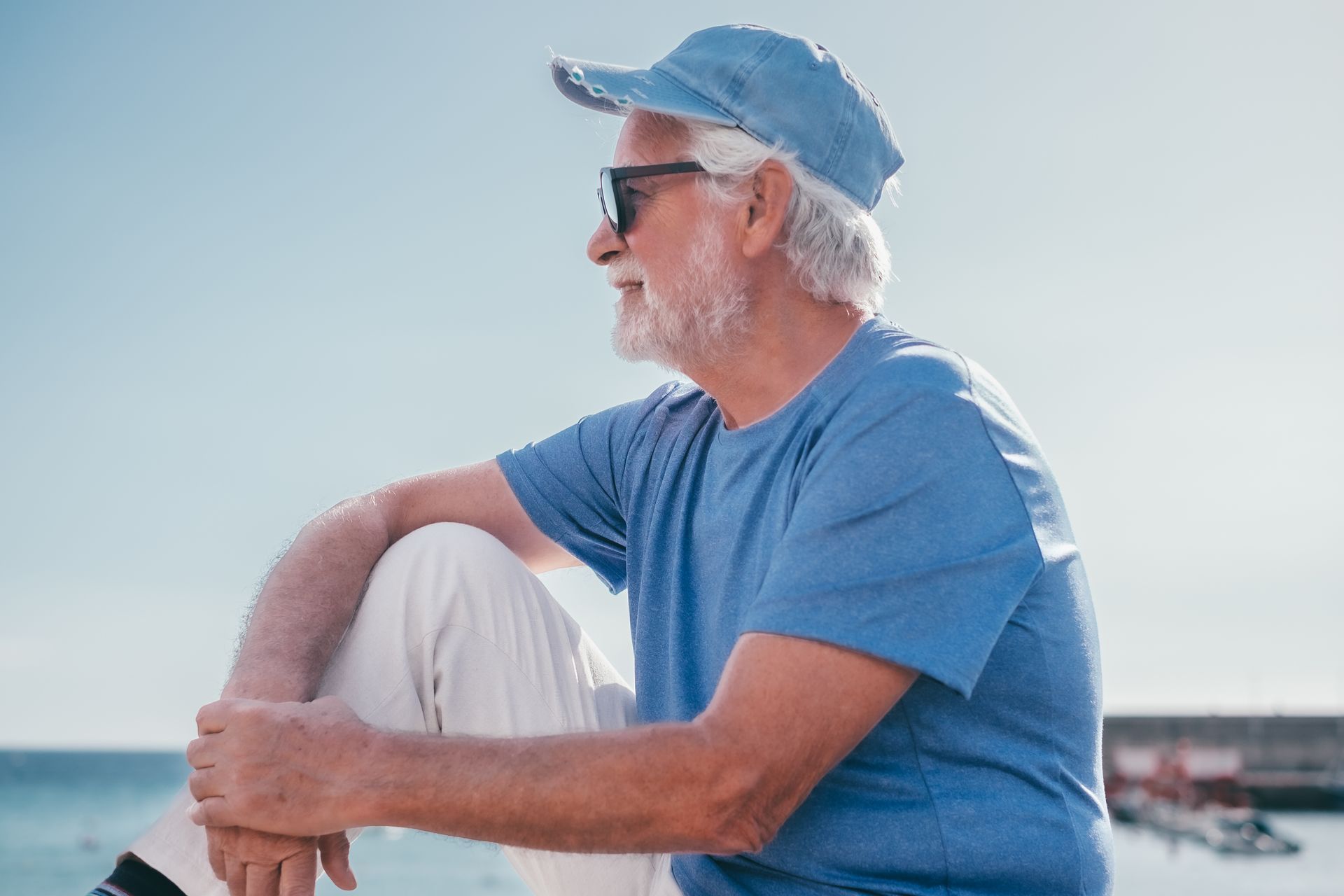 Elderly man in blue shirt and hat, sunglasses, looks out at the ocean.