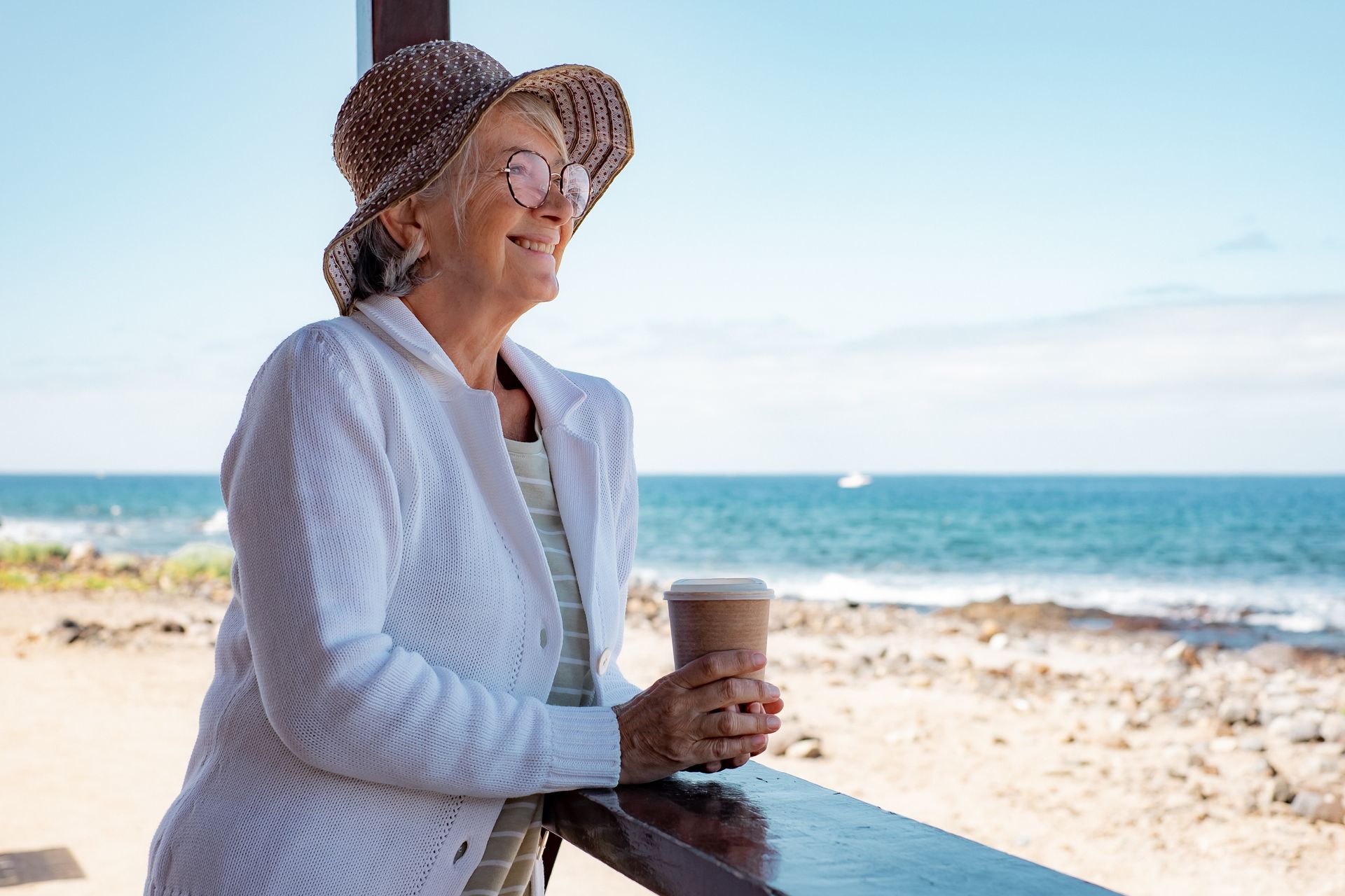 Older woman in a hat and sweater smiles, holding a coffee, looking out at the ocean.