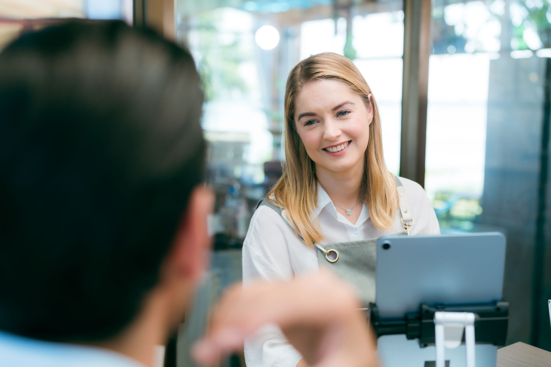 Woman taking order behind counter, smiling at customer. Cafe setting, light-colored clothing.