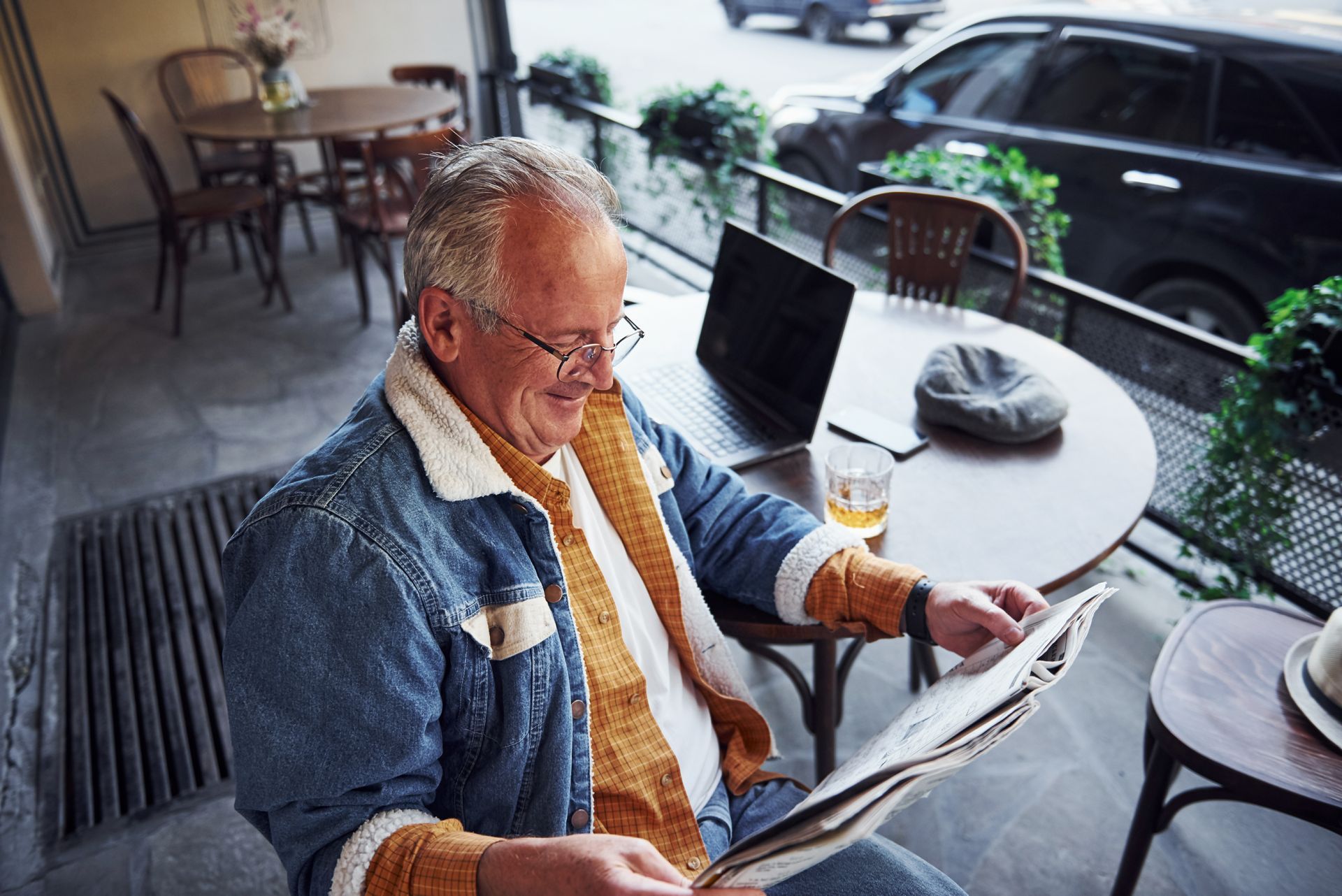 Man wearing glasses and denim jacket reading a newspaper, sitting at a cafe table with laptop and drink.