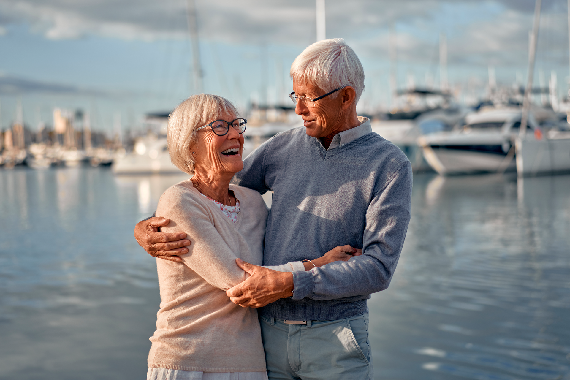 Smiling elderly couple embracing by the water, boats in background.