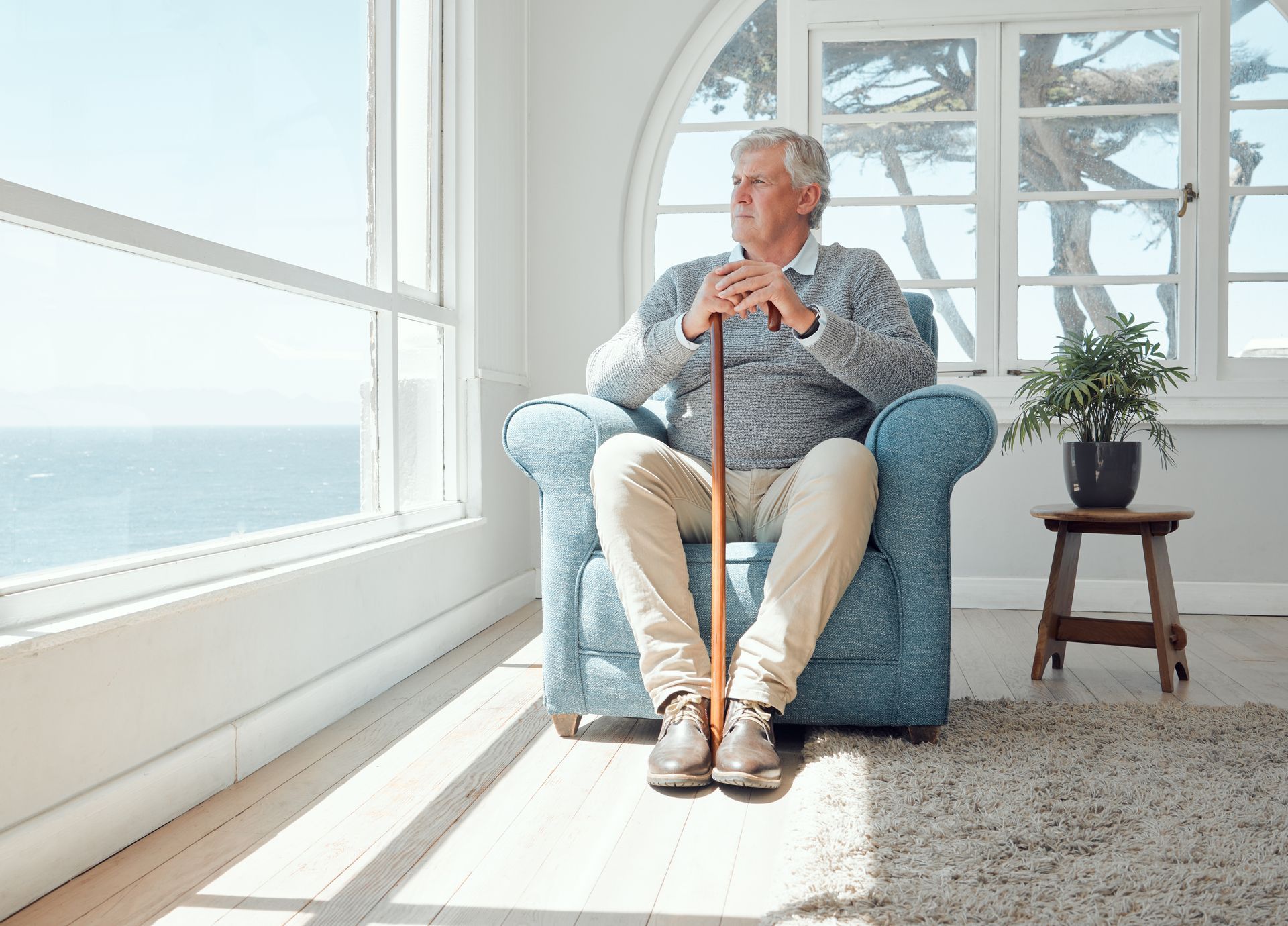 Elderly man sits in a blue armchair by a window, holding a cane, looking out at the ocean.