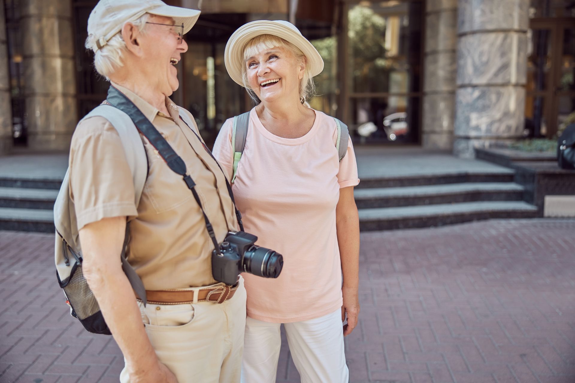 Elderly couple laughing together outside a building, both wearing hats and carrying cameras.