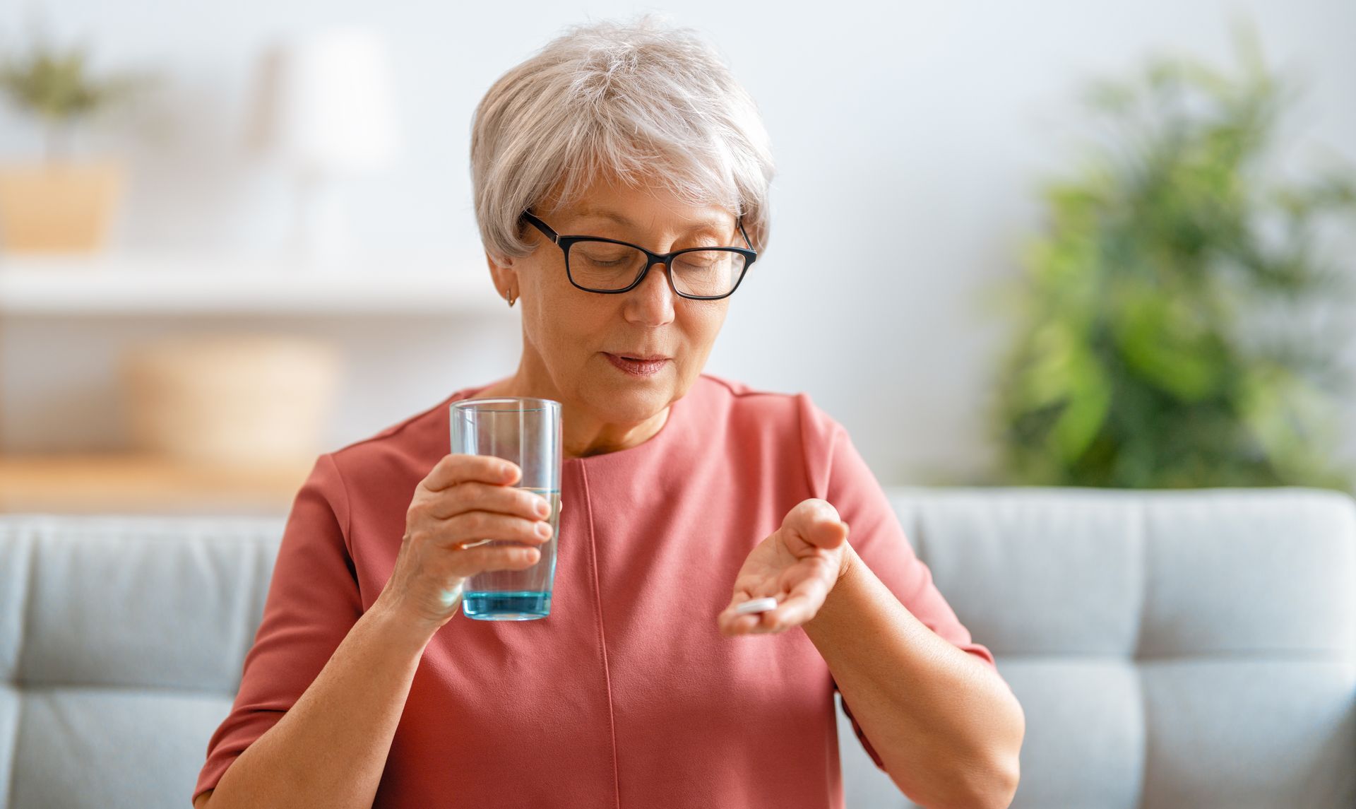 Woman taking pills, holding a glass of water, sitting on a couch.