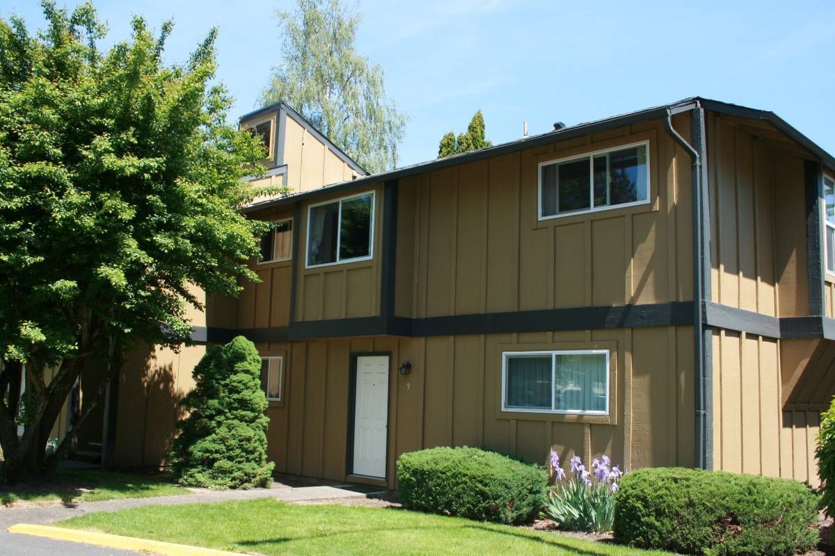 Two-story tan apartment building with white-framed windows, surrounded by greenery, on a sunny day.