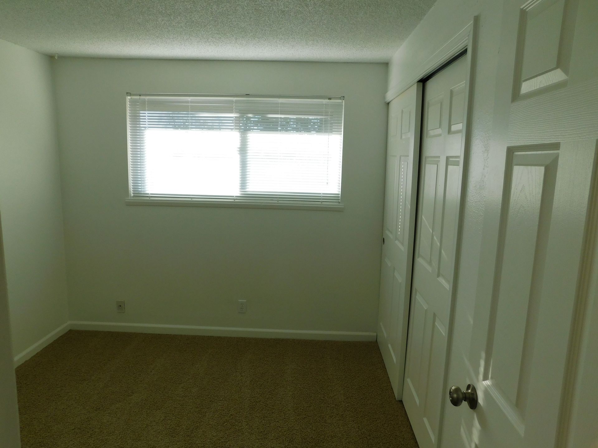 Empty bedroom with brown carpet, white walls, window with blinds, and sliding closet doors.