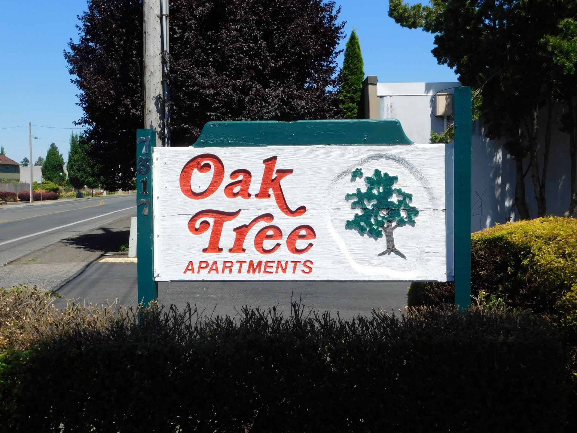 Sign for Oak Tree Apartments: white with red text, green oak tree illustration, street in background.