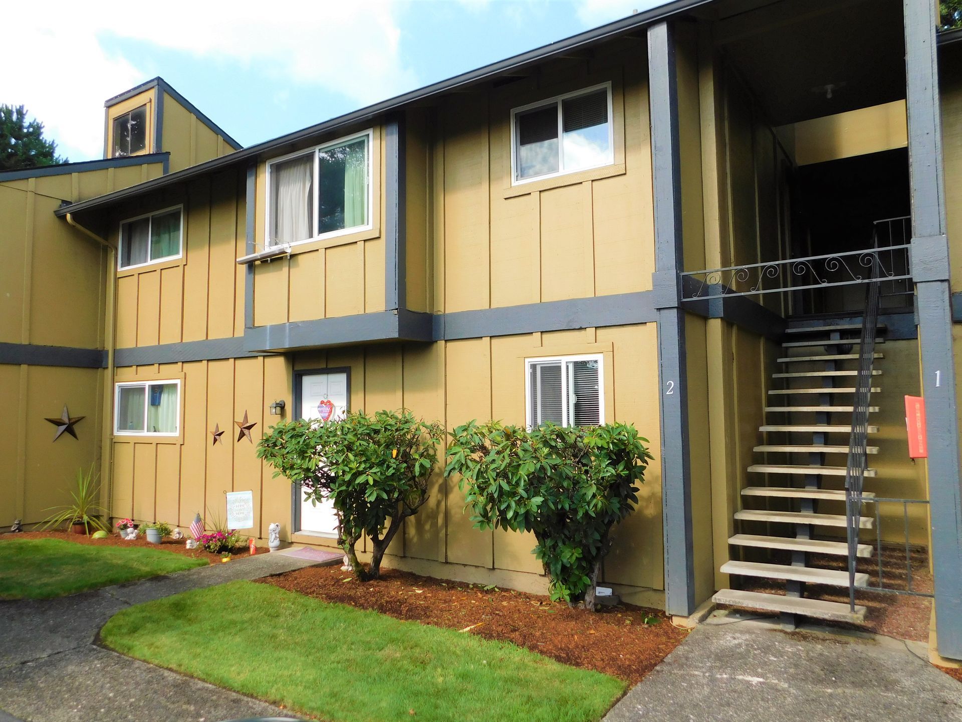 Two-story tan apartment building with large tree in front; sunny day.