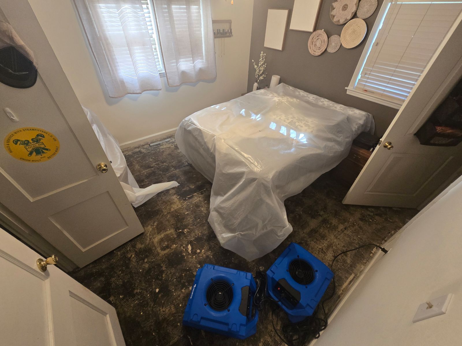 A bedroom with a bed wrapped in plastic and two fans.