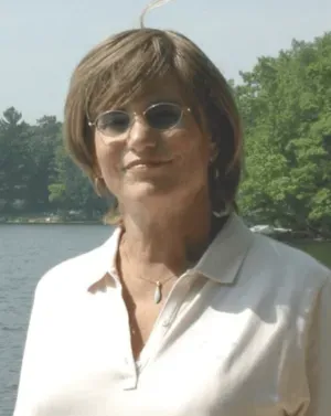 Woman with short brown hair and sunglasses, smiles slightly. She wears a light-colored shirt, outdoors by a lake.