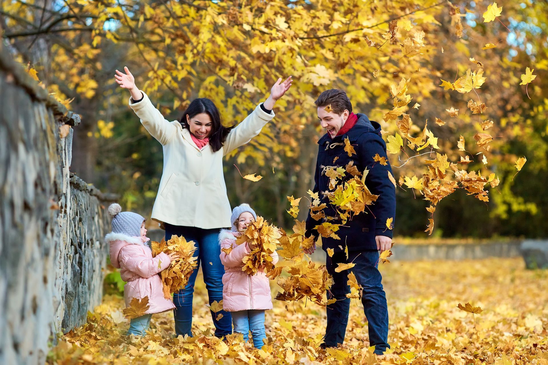 Family throws autumn leaves in a park; woman with arms up, children in pink coats.