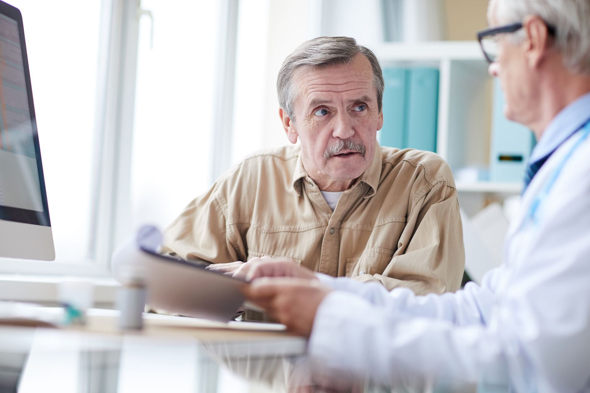 Man in tan shirt talking to doctor, who holds a clipboard, in a doctor's office.
