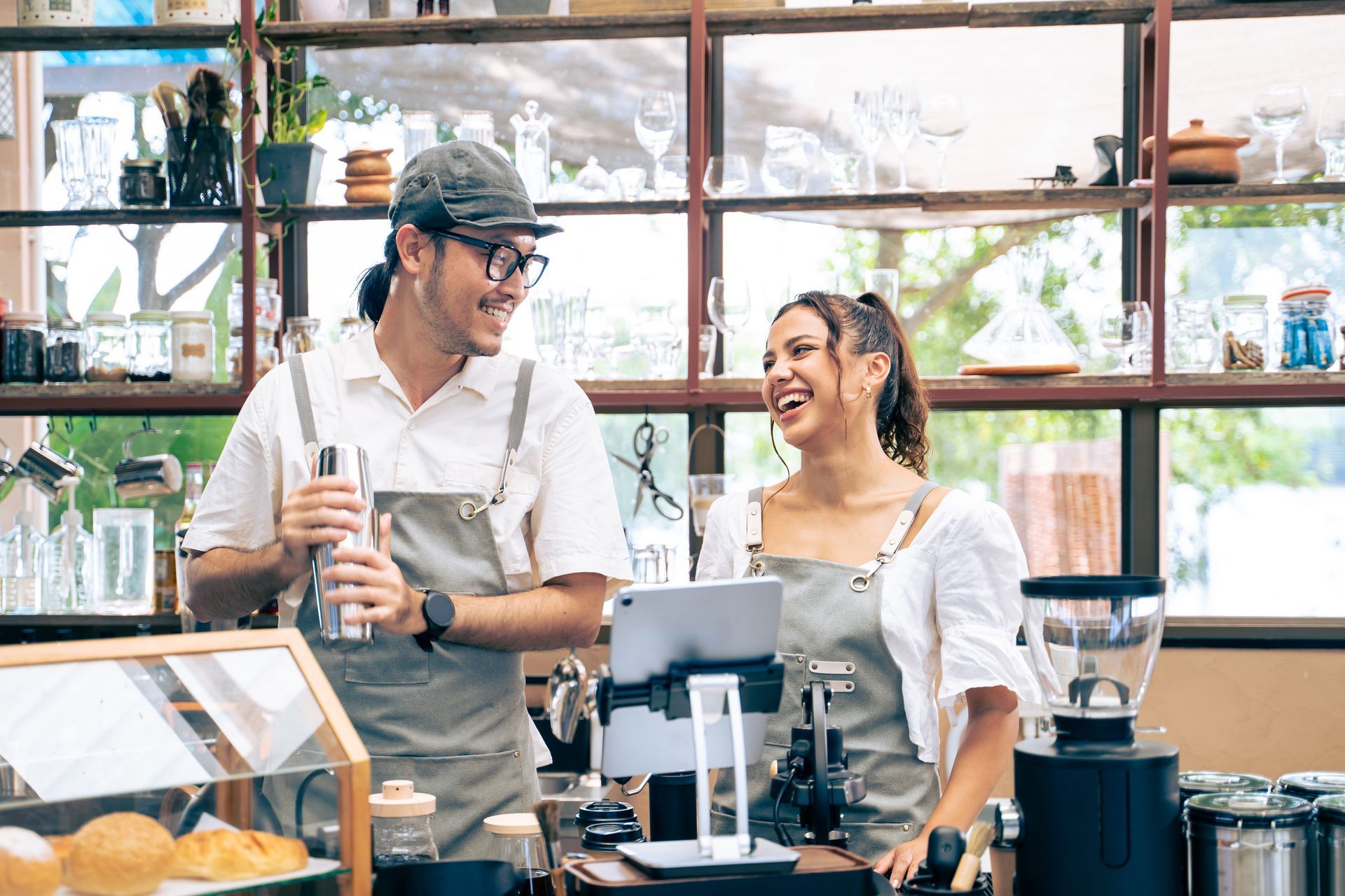 Two baristas laugh while working in a cafe. One holds a shaker, the other looks at a tablet.