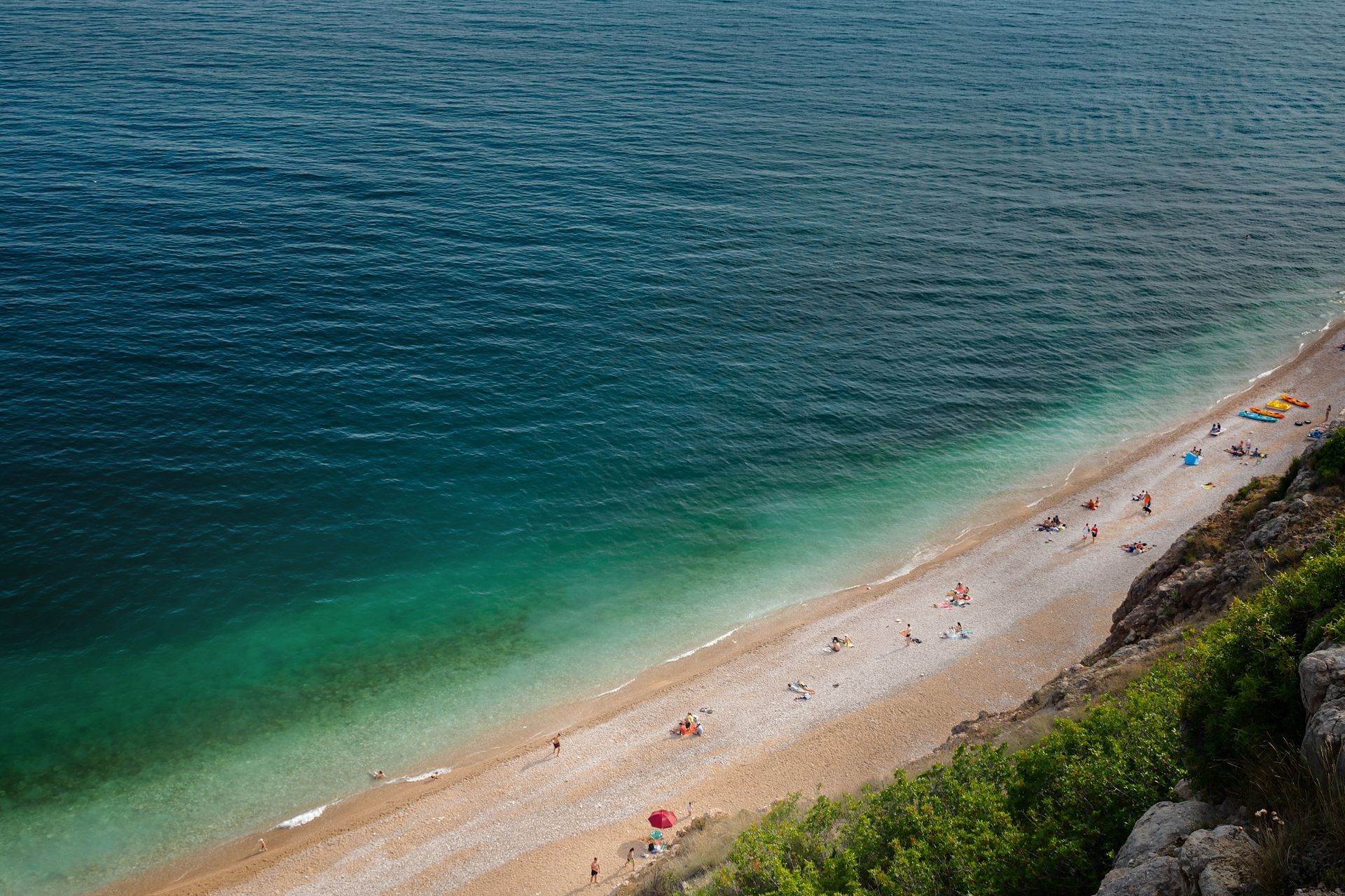 Beach with turquoise water, rocky shore, people sunbathing. View from cliff with green foliage.