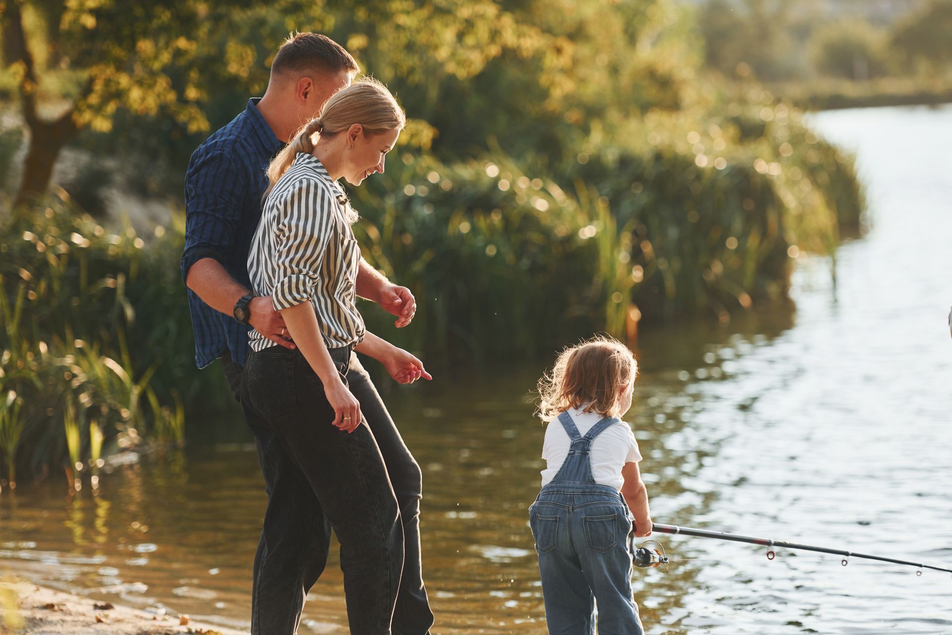 Family by a lake fishing. Parents watch child in overalls holding a fishing rod.