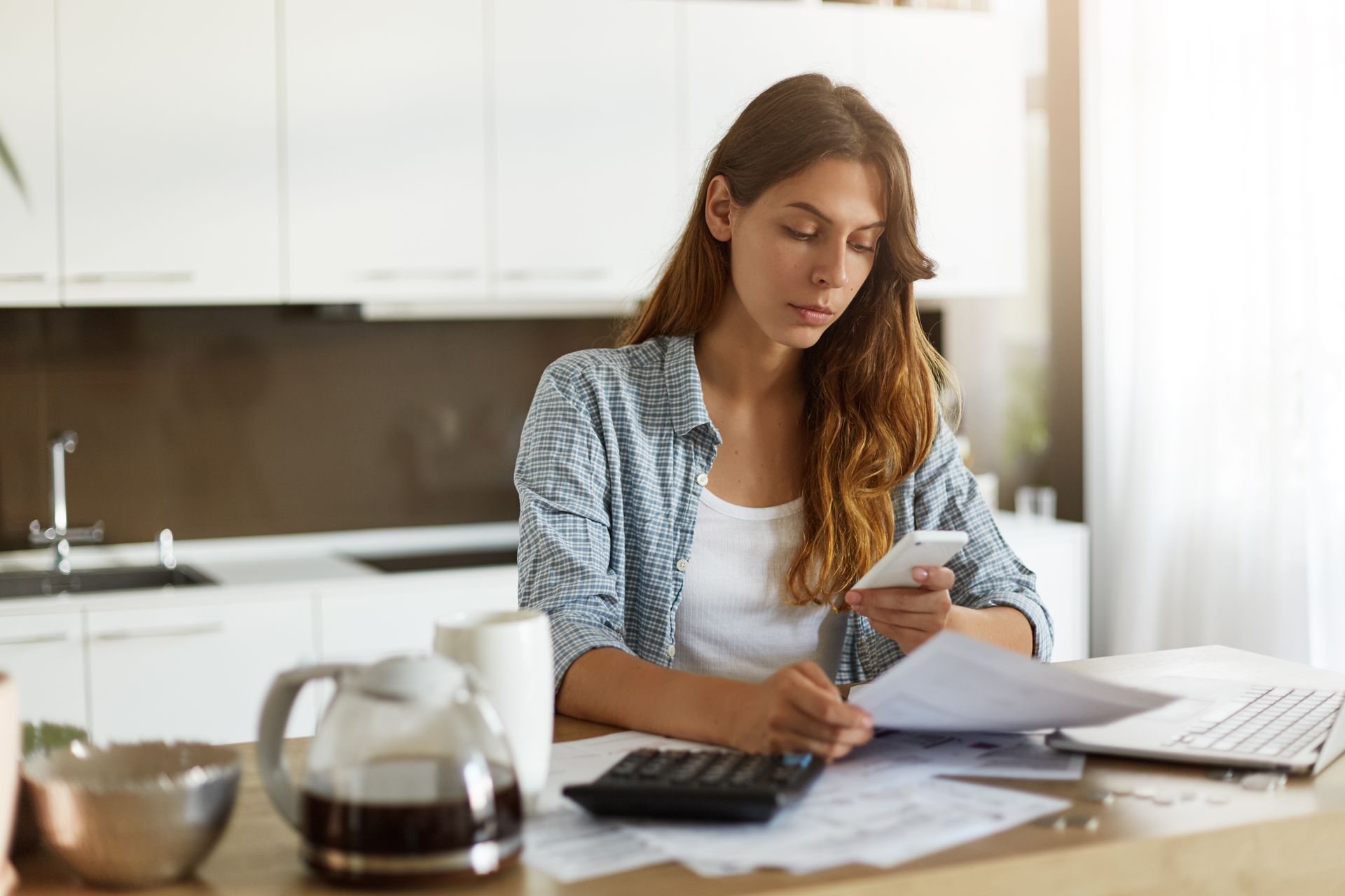 Woman in kitchen, using phone while reviewing documents and calculating with a calculator.