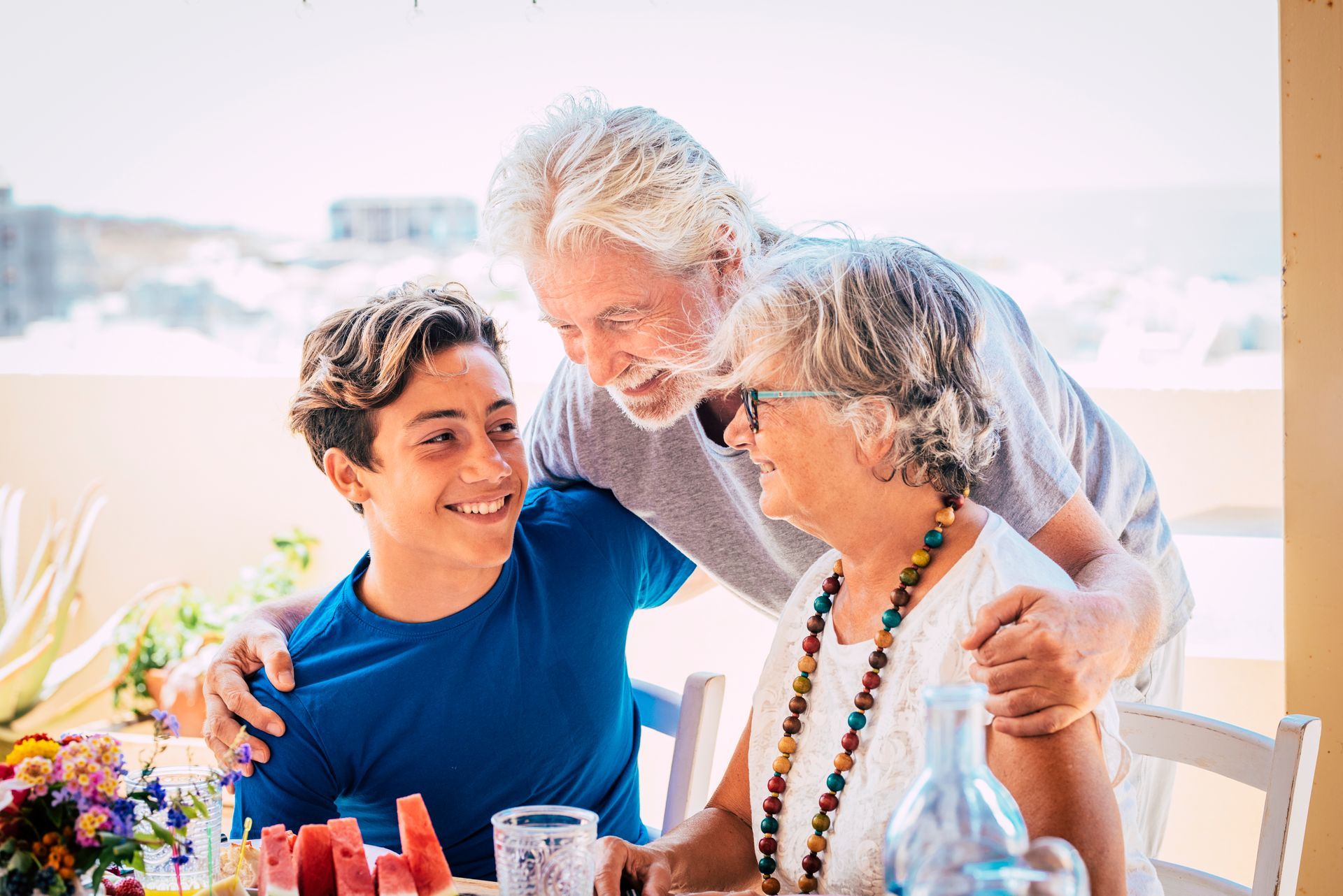 Grandparents embrace a smiling teen at a table with food, outdoors.