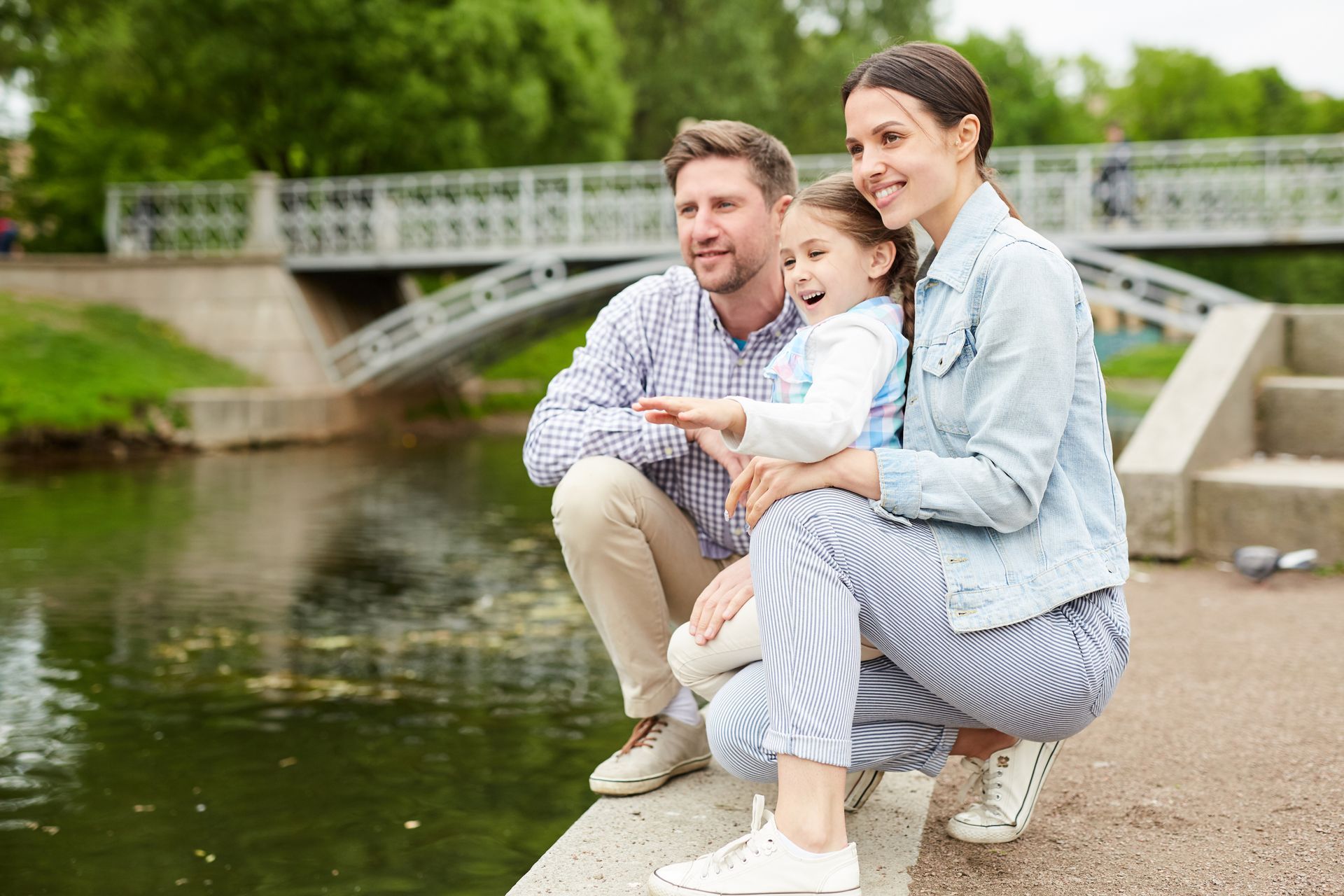Family of three by a pond, pointing, smiling, bridge in background.