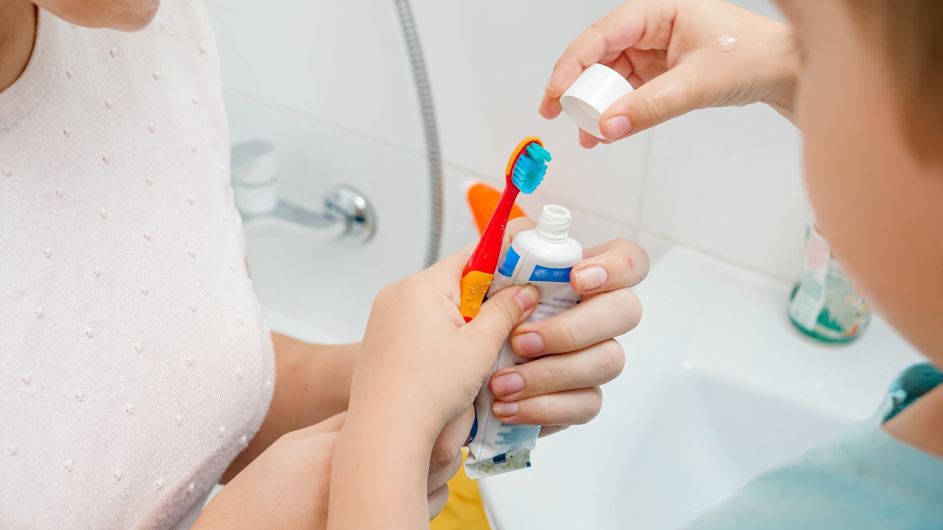 Woman squeezing toothpaste onto child's toothbrush in bathroom with sink and faucet.