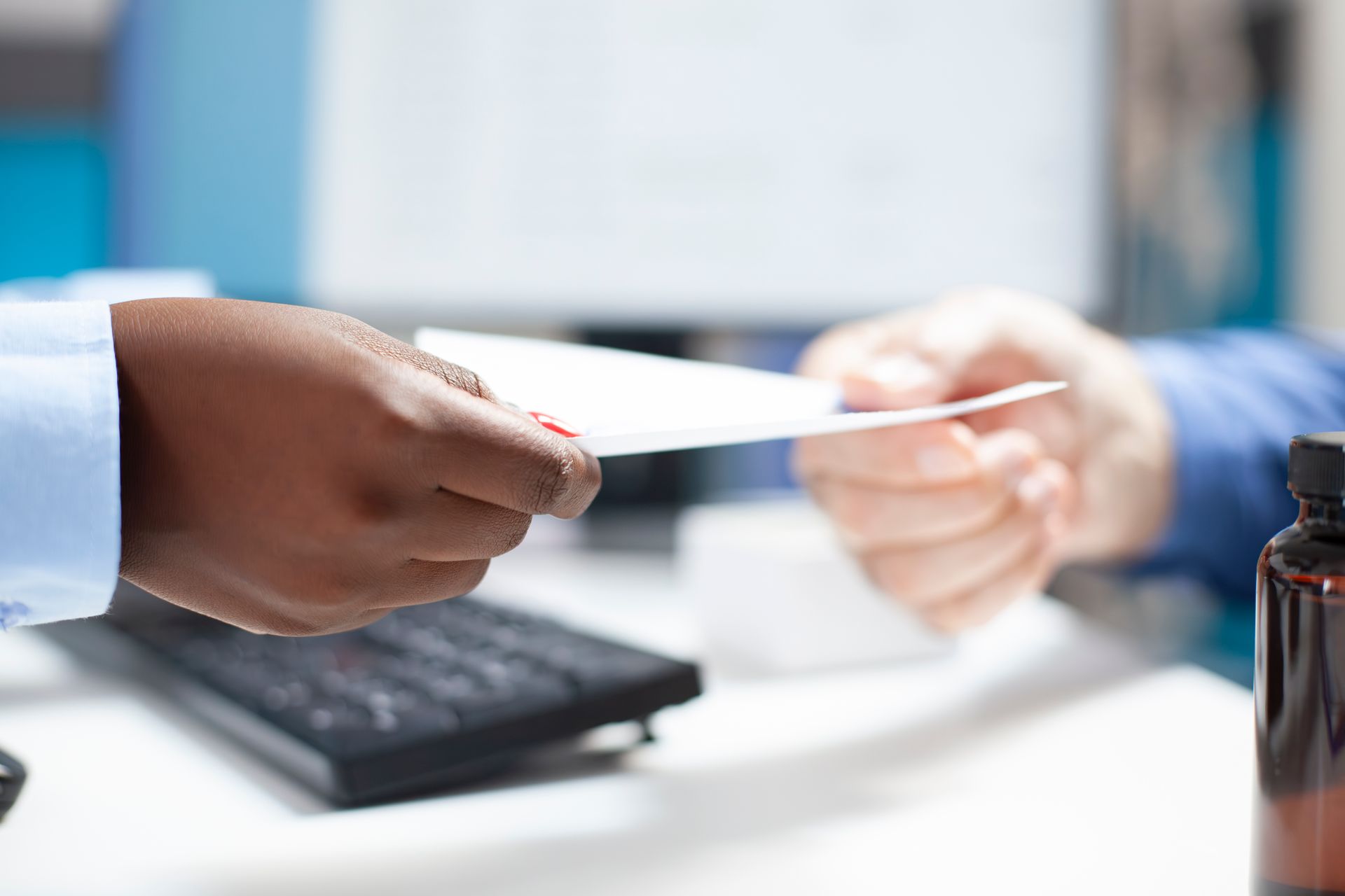 Hands exchanging a white paper; a keyboard and brown bottle are nearby.