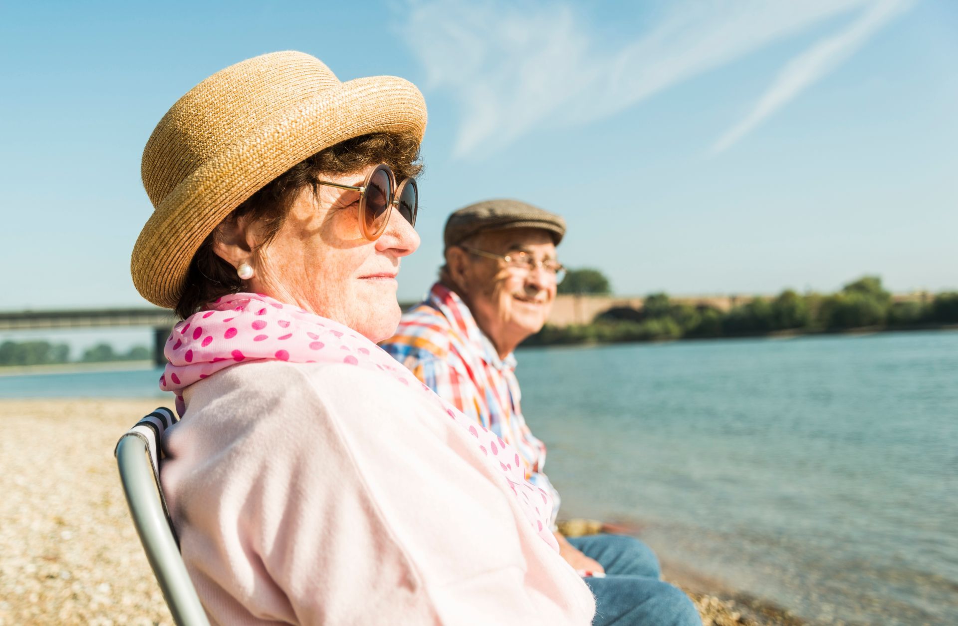 Elderly couple seated by water, woman wearing hat and sunglasses, man in cap, sunny day.