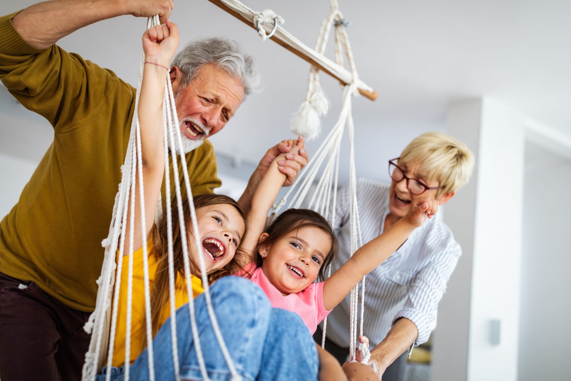 Grandparents push two laughing children on a swing indoors.