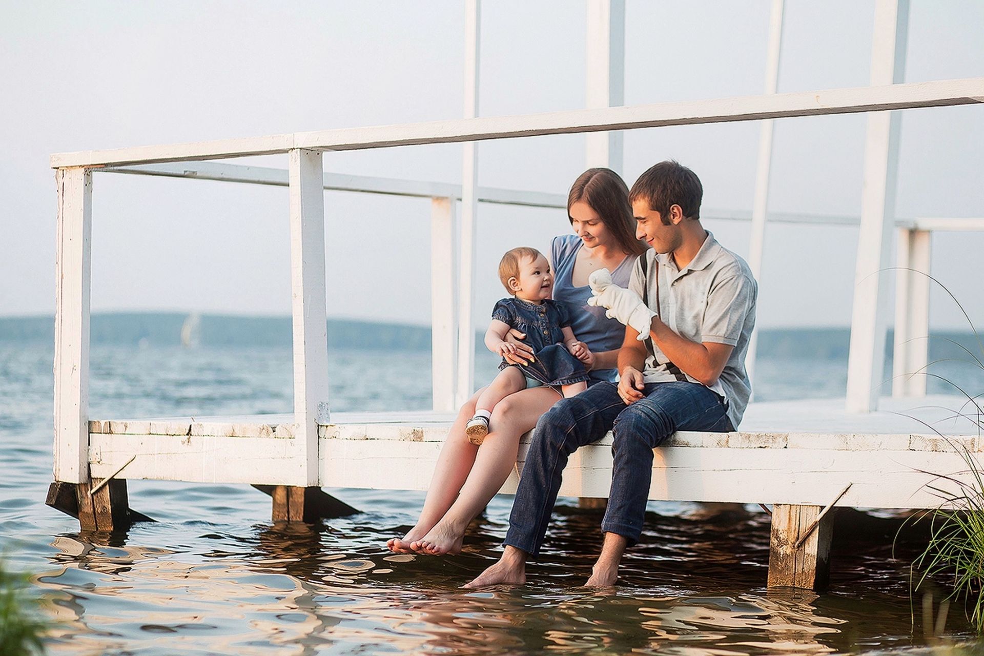 Family of three on white pier, baby smiling, near water.