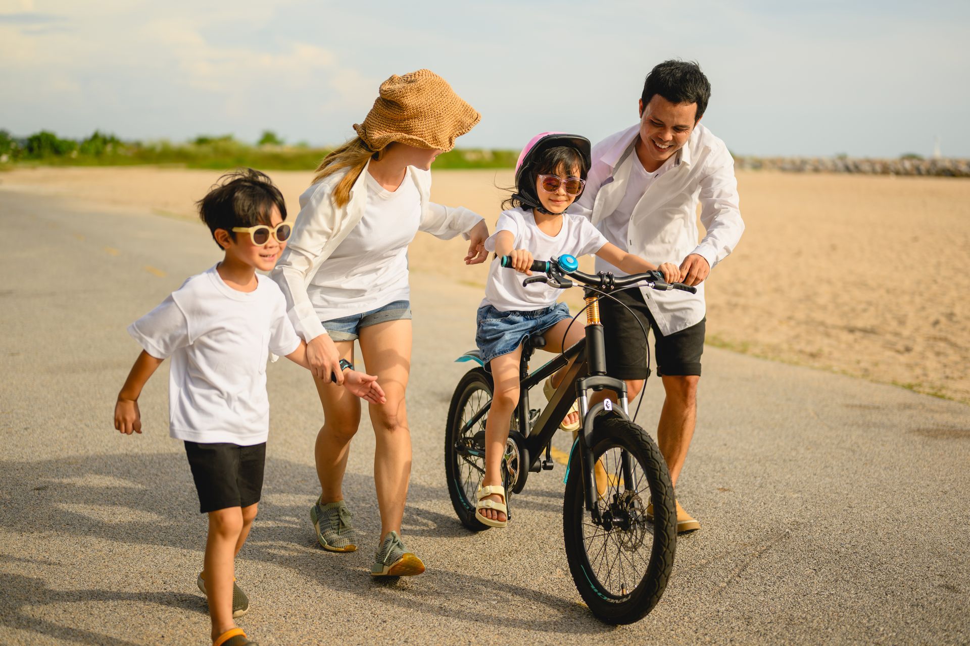 Family helps a girl learn to ride a bike on a paved path.
