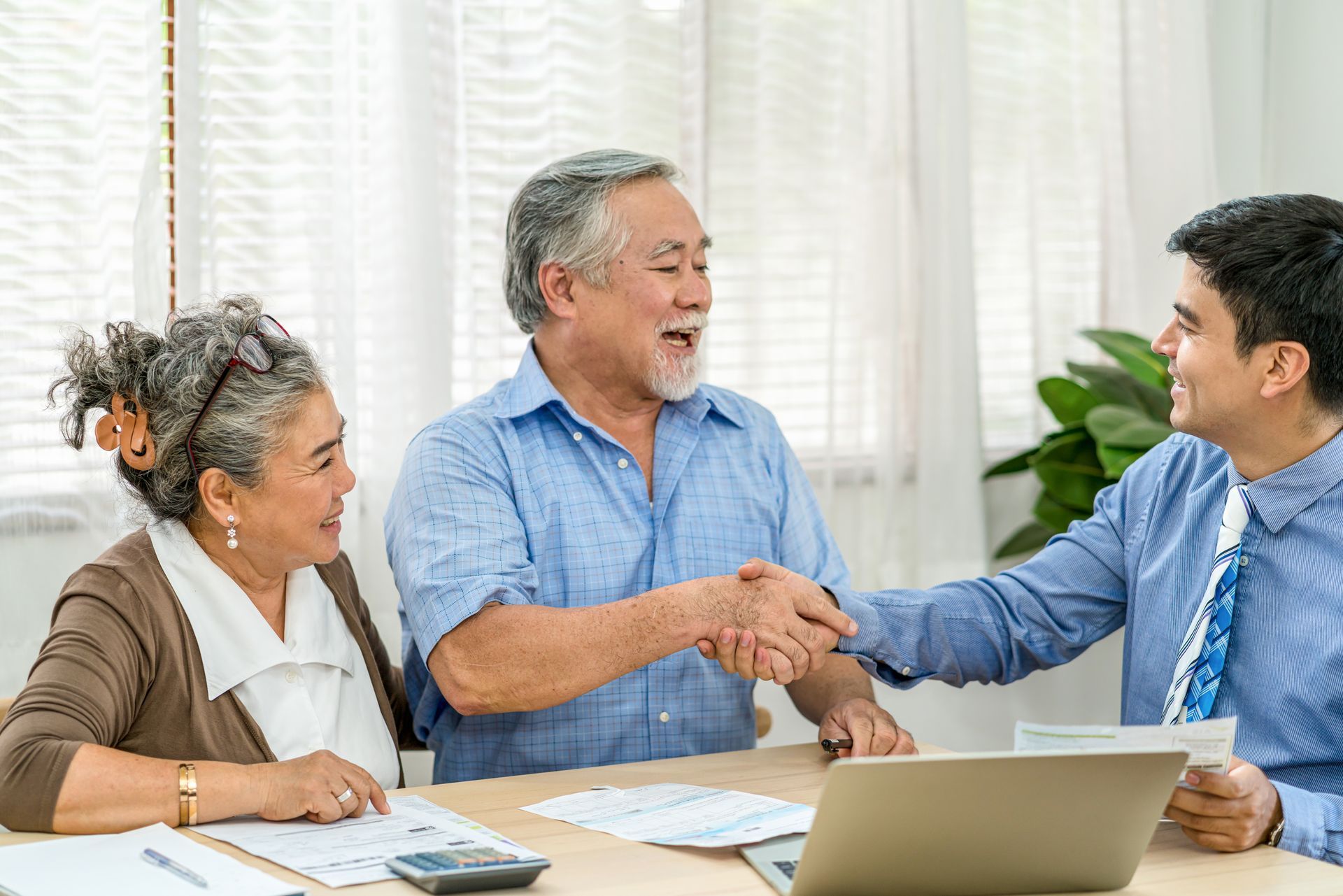 Elderly couple shakes hands with a professional at a table with papers, calculator, and laptop.
