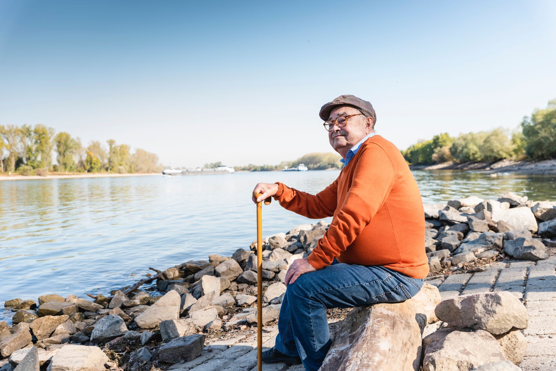 Man in orange sweater and cap sits on a rock by a river, holding a cane, looking at the camera.