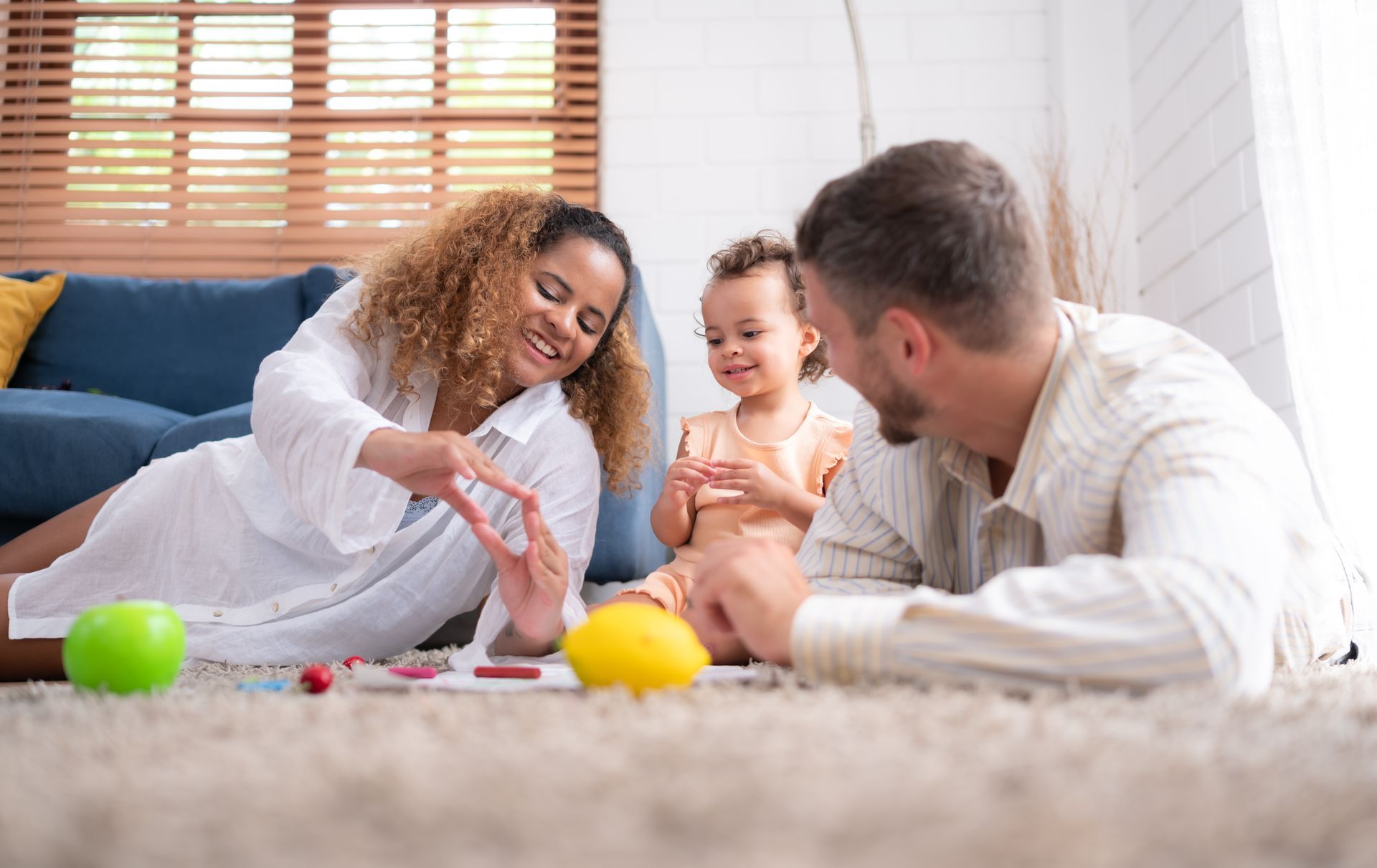 Family playing on a rug. The woman makes a heart with her hands. Child and man watch, smiling. Bright room.