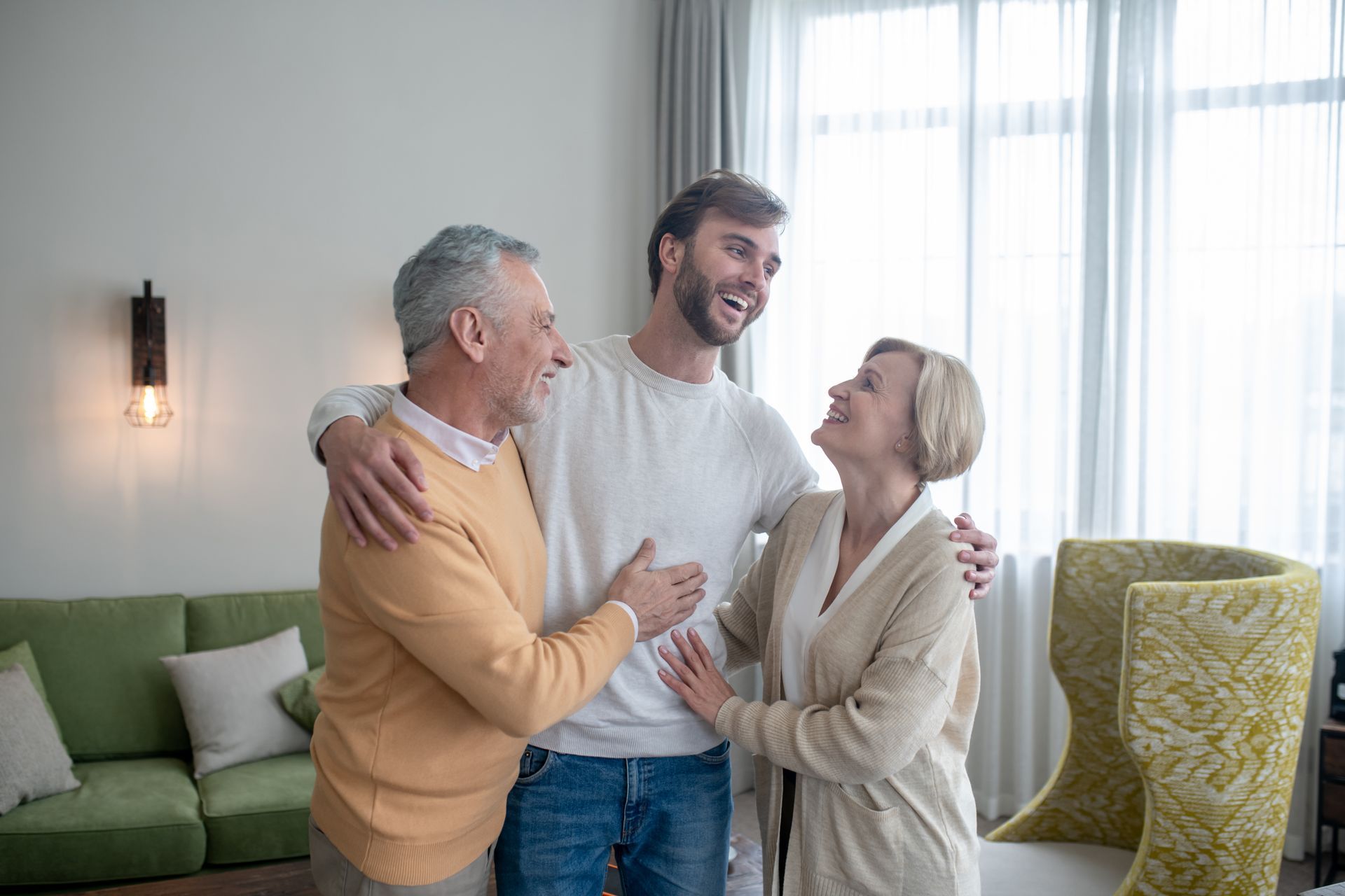 Man hugs older couple; they smile in a bright room with a window, couch, and chair.