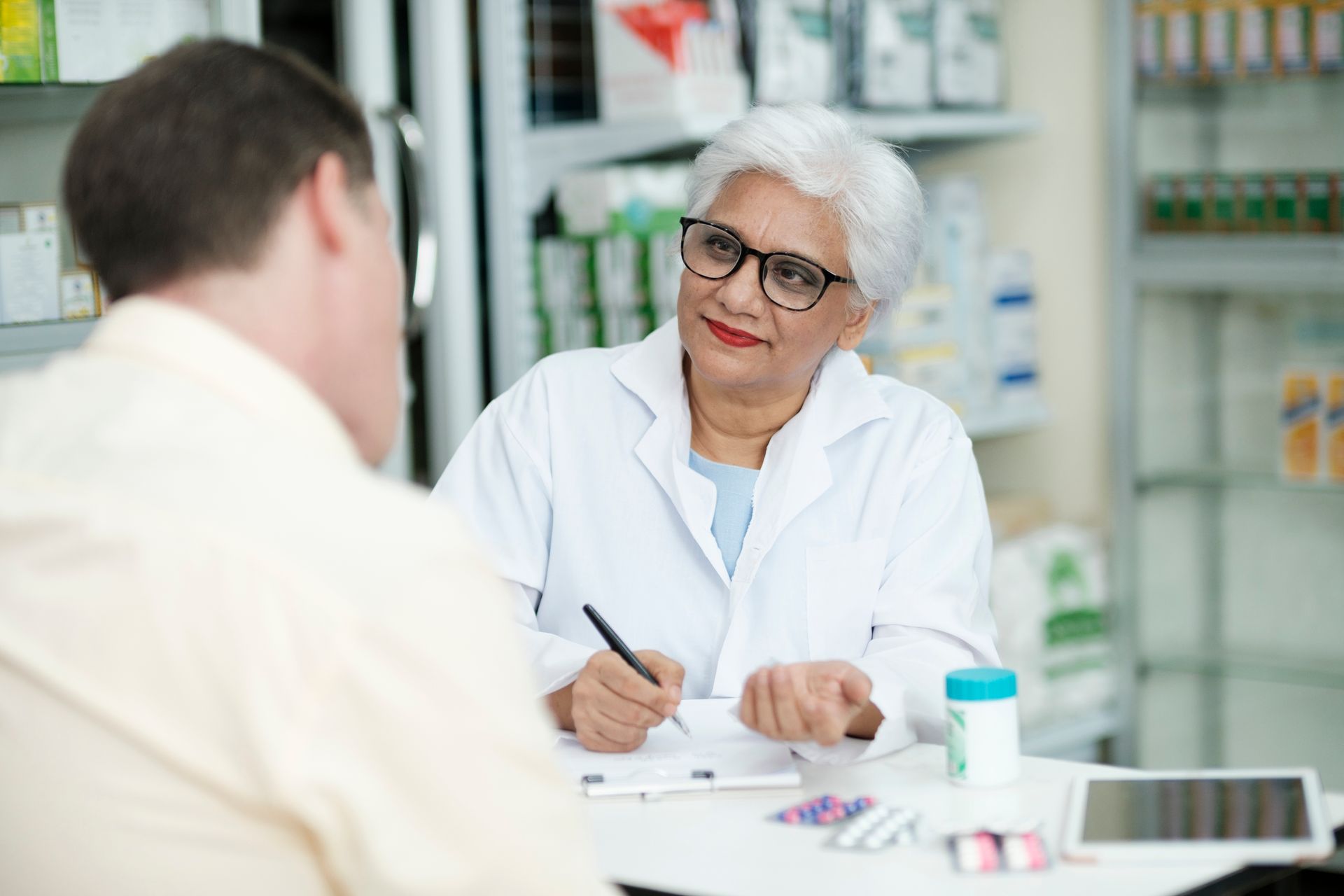 Pharmacist writing notes while talking to a customer at a pharmacy counter.