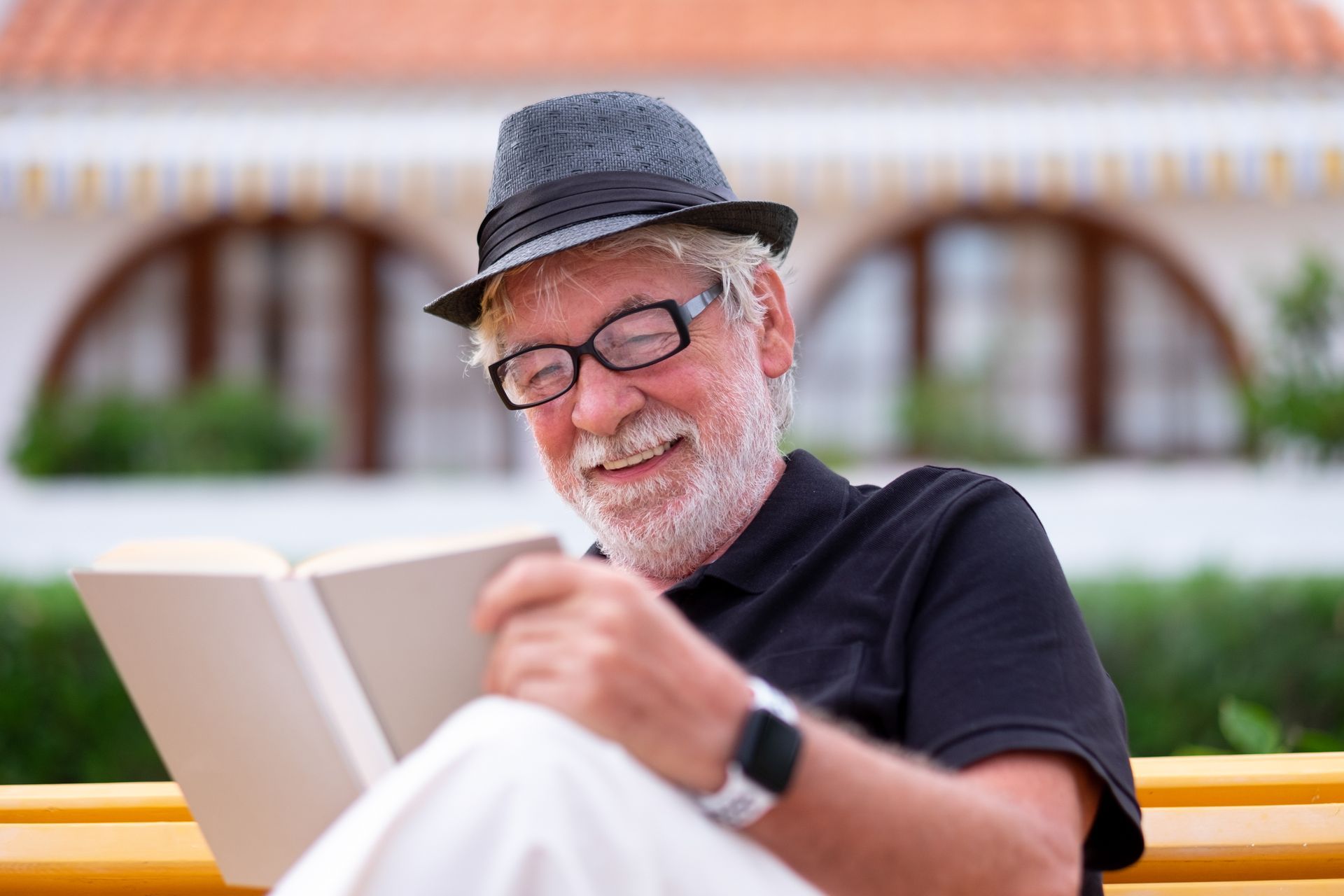 Man in hat and glasses smiles while reading a book on a bench.