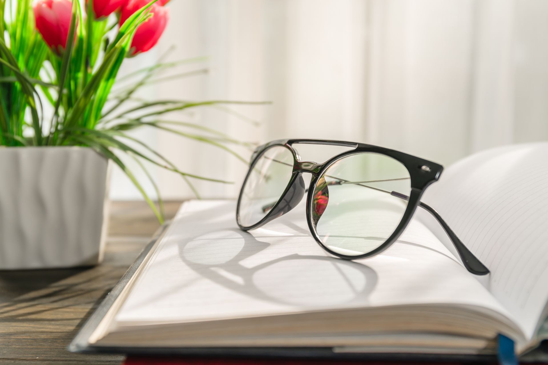 Black-framed glasses rest on an open book, next to a vase with red flowers; daylight from a window illuminates the scene.