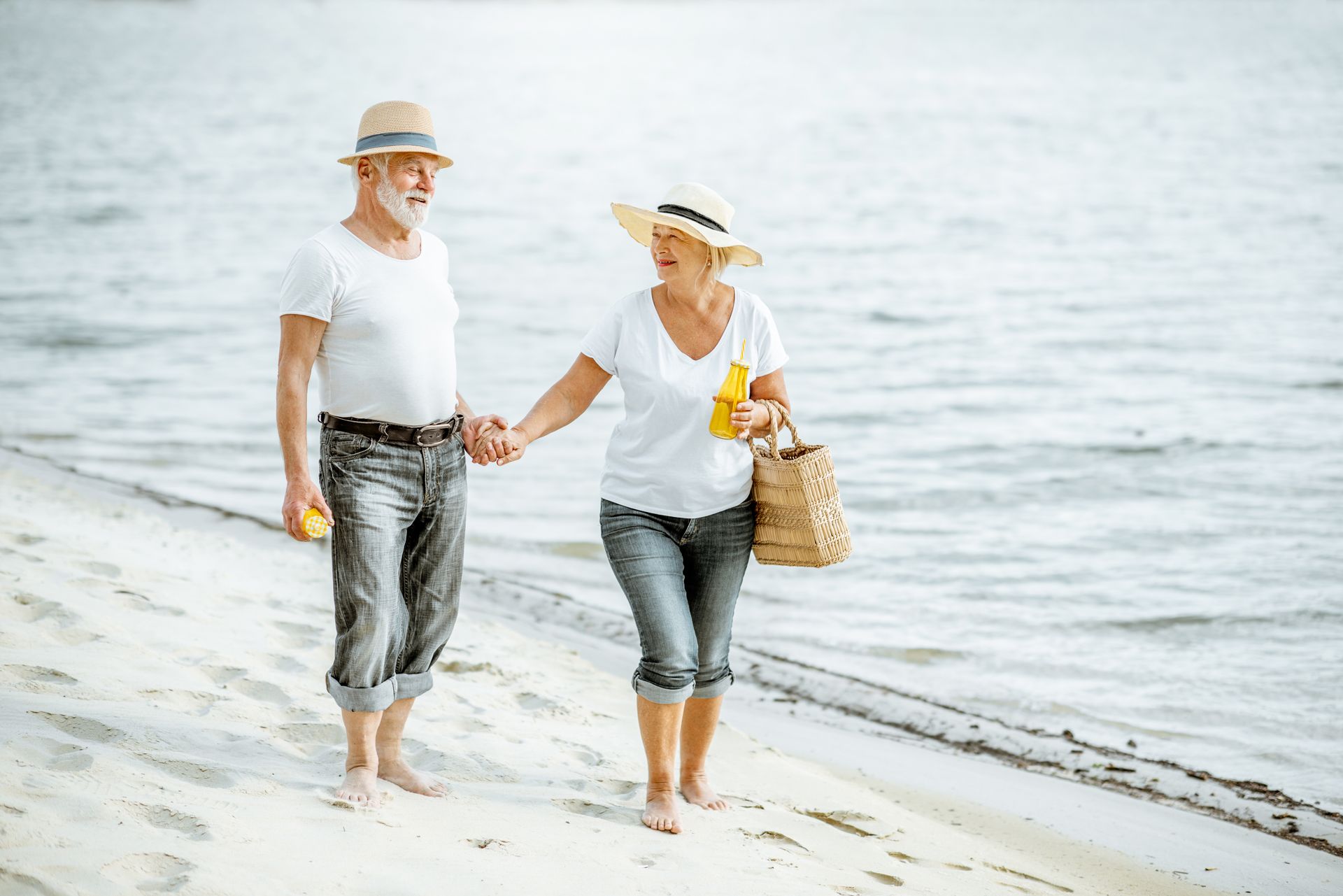 Elderly couple holding hands, walking on a beach, near water. Both are wearing hats and white shirts.