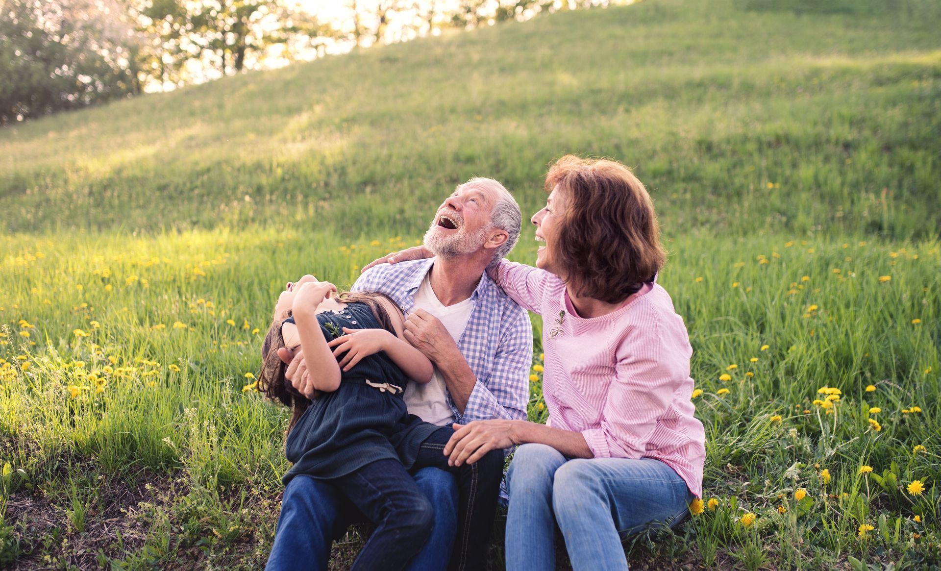 Grandparents and child laughing together in a sunny field of green grass and yellow flowers.