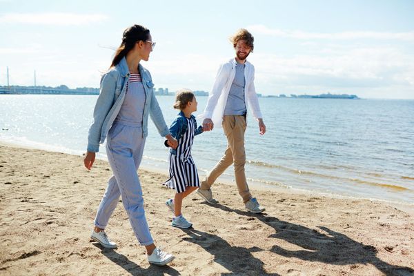 Family of three walking on a sandy beach holding hands, sunny day.