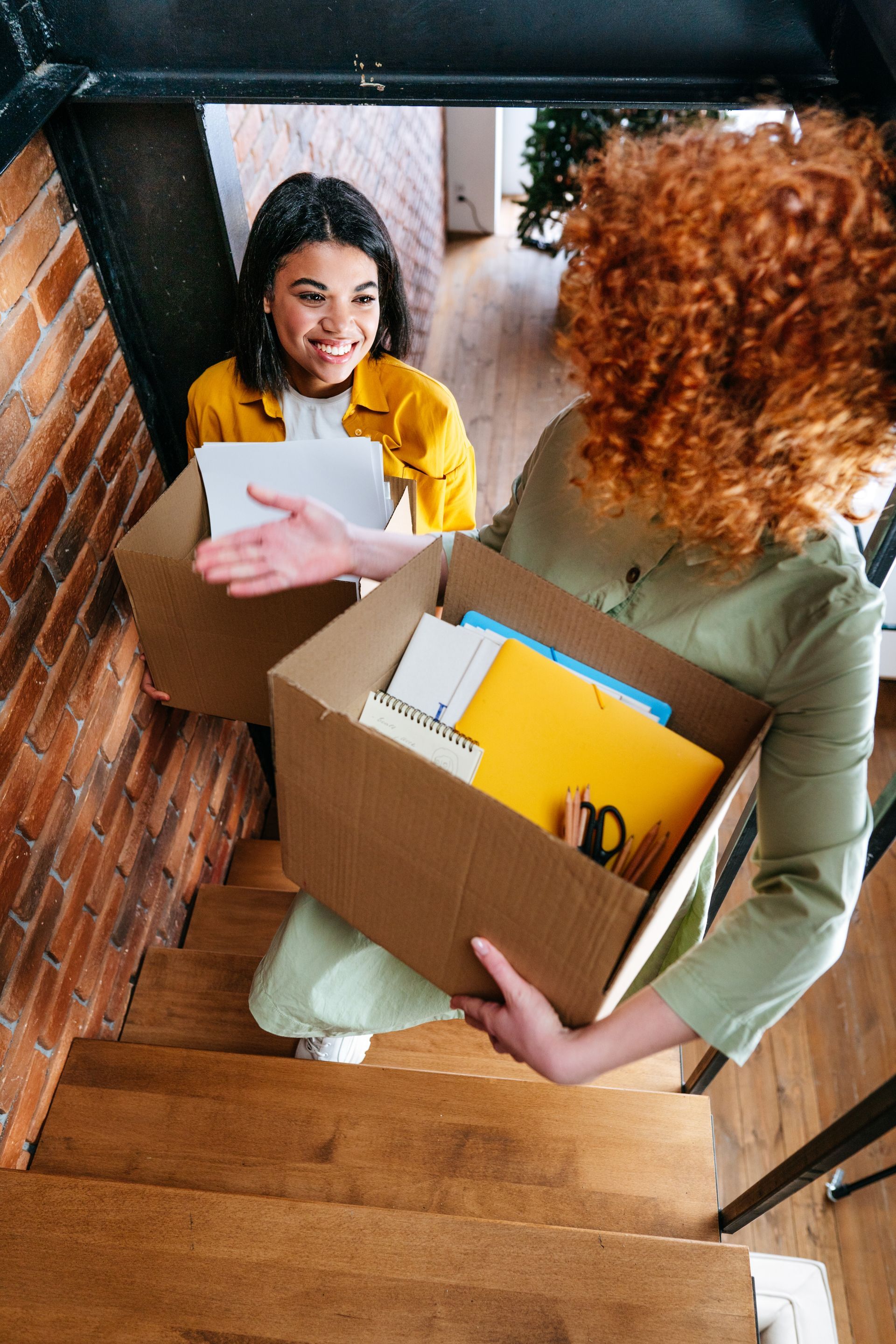 Two women moving boxes on a staircase. One smiles, the other carries a box, ascending. Red brick wall.