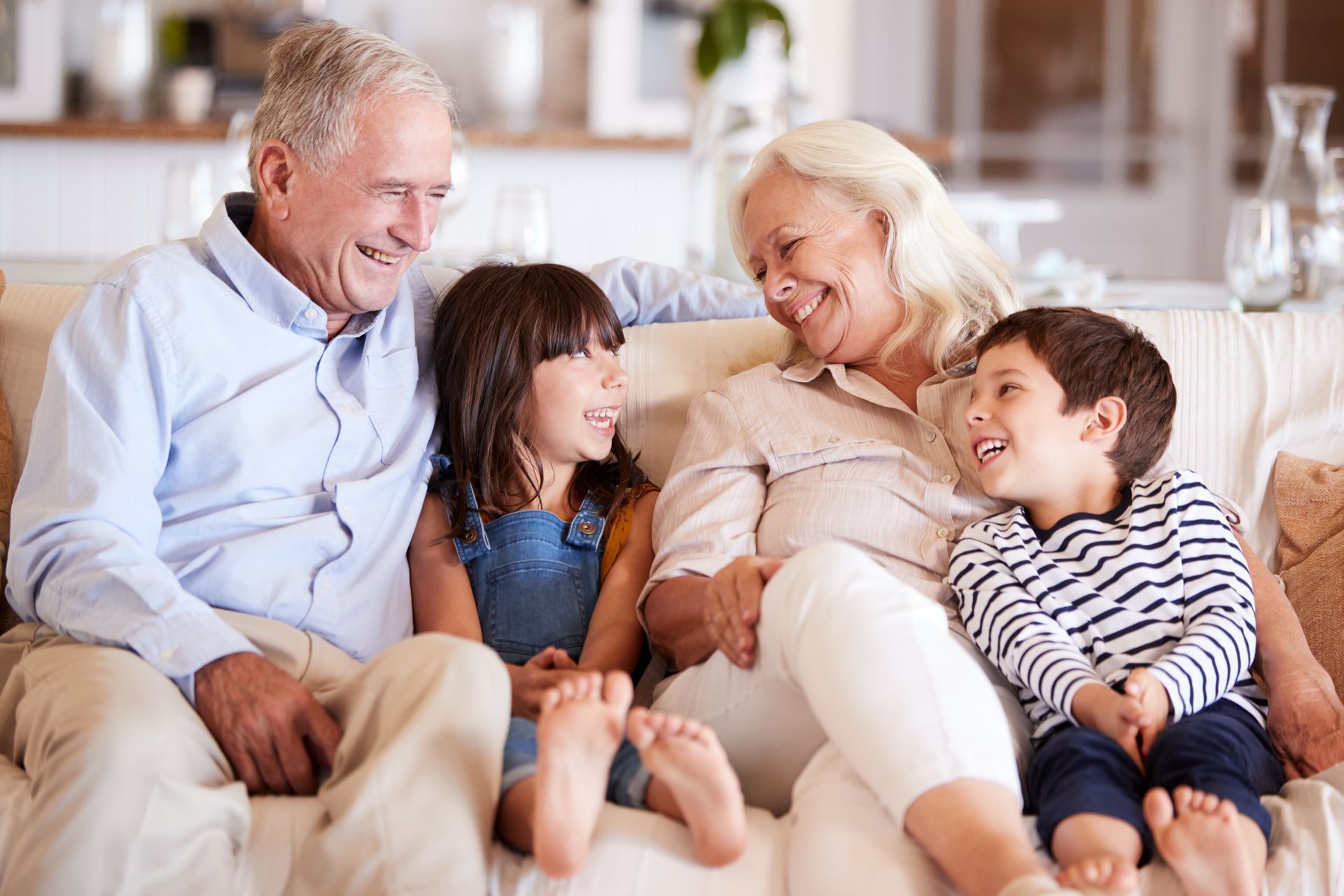 Grandparents smiling with two grandchildren on a beige sofa indoors.