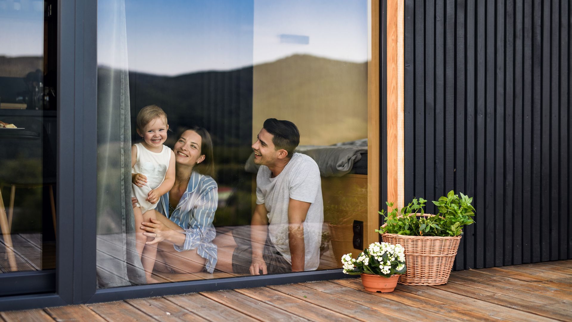 Family smiling through a large window, inside a modern cabin with wooden siding.