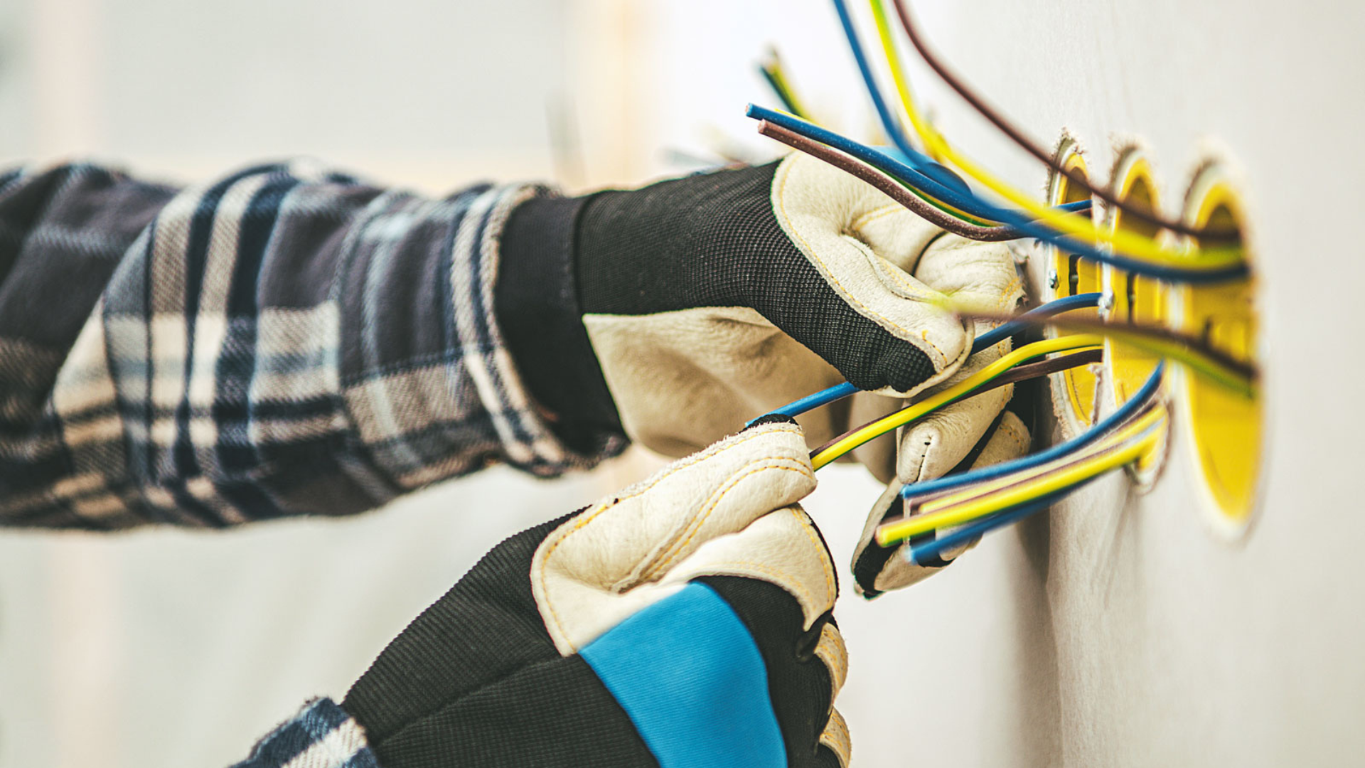 Electrician's hands, wearing gloves, work with colorful electrical wires in a wall outlet.
